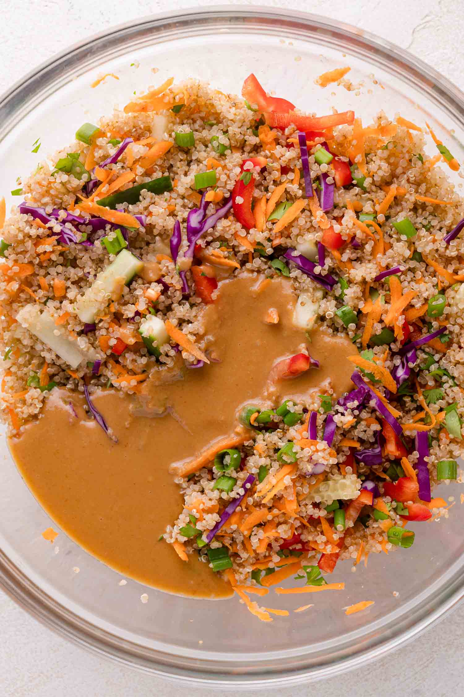 A glass bowl filled with quinoa, chopped vegetables, and shredded carrots, topped with a thick brown dressing.