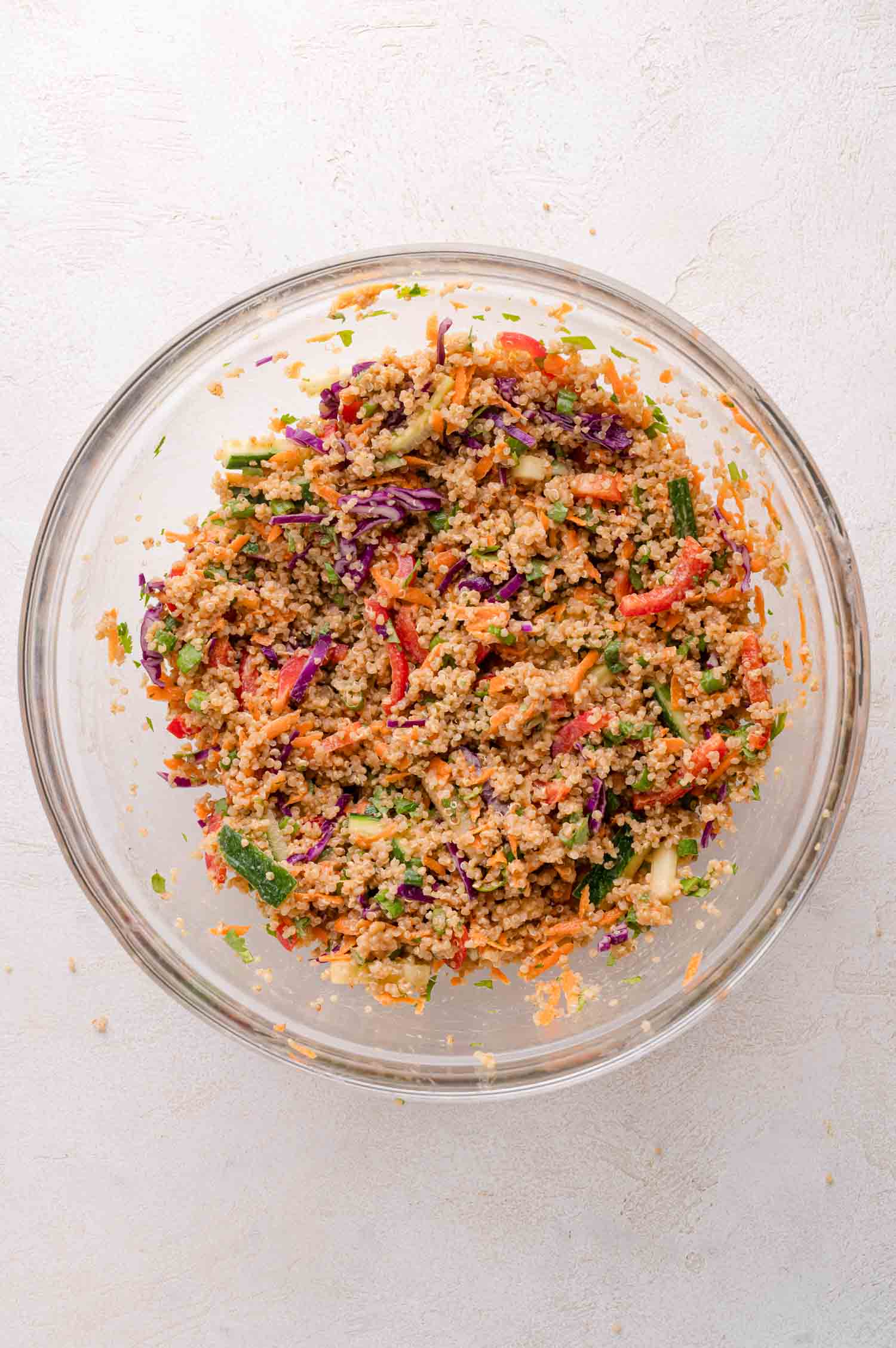 A glass bowl filled with a colorful quinoa salad containing shredded carrots, red cabbage, bell peppers, cucumbers, and herbs on a light surface.