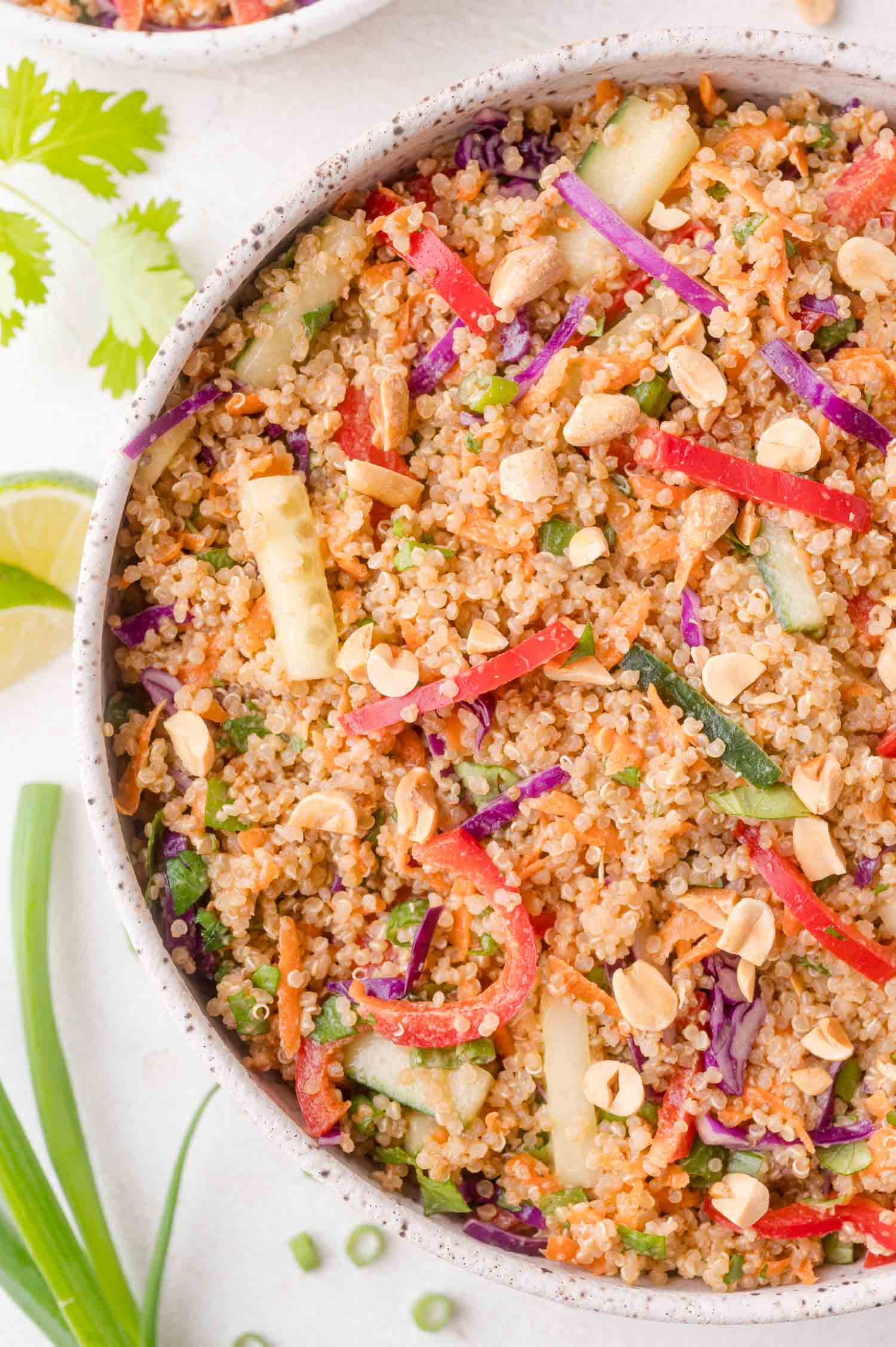 A bowl of quinoa spring roll salad with chopped red bell peppers, cucumbers, shredded carrots, purple cabbage, green onions, and crushed peanuts.