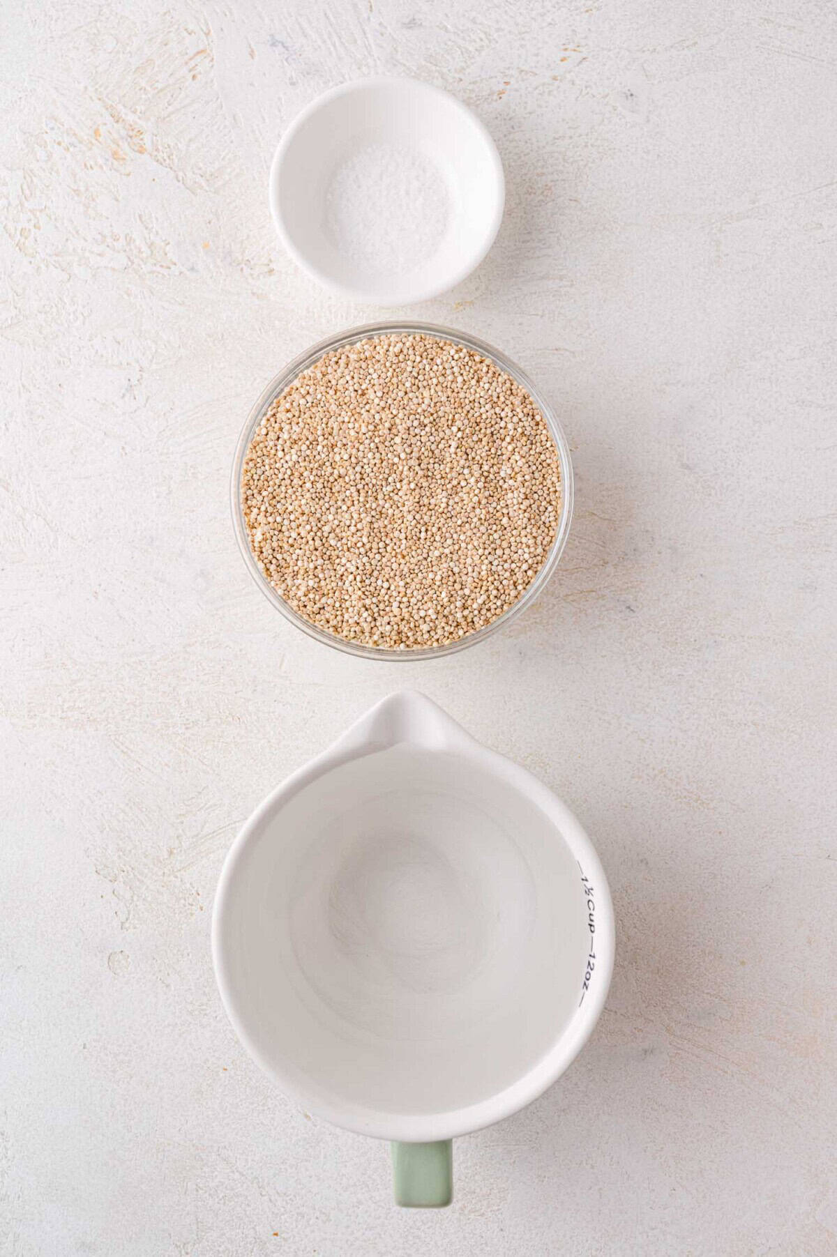 A small bowl of salt, a bowl of uncooked quinoa, and an empty measuring cup are arranged vertically on a light surface.