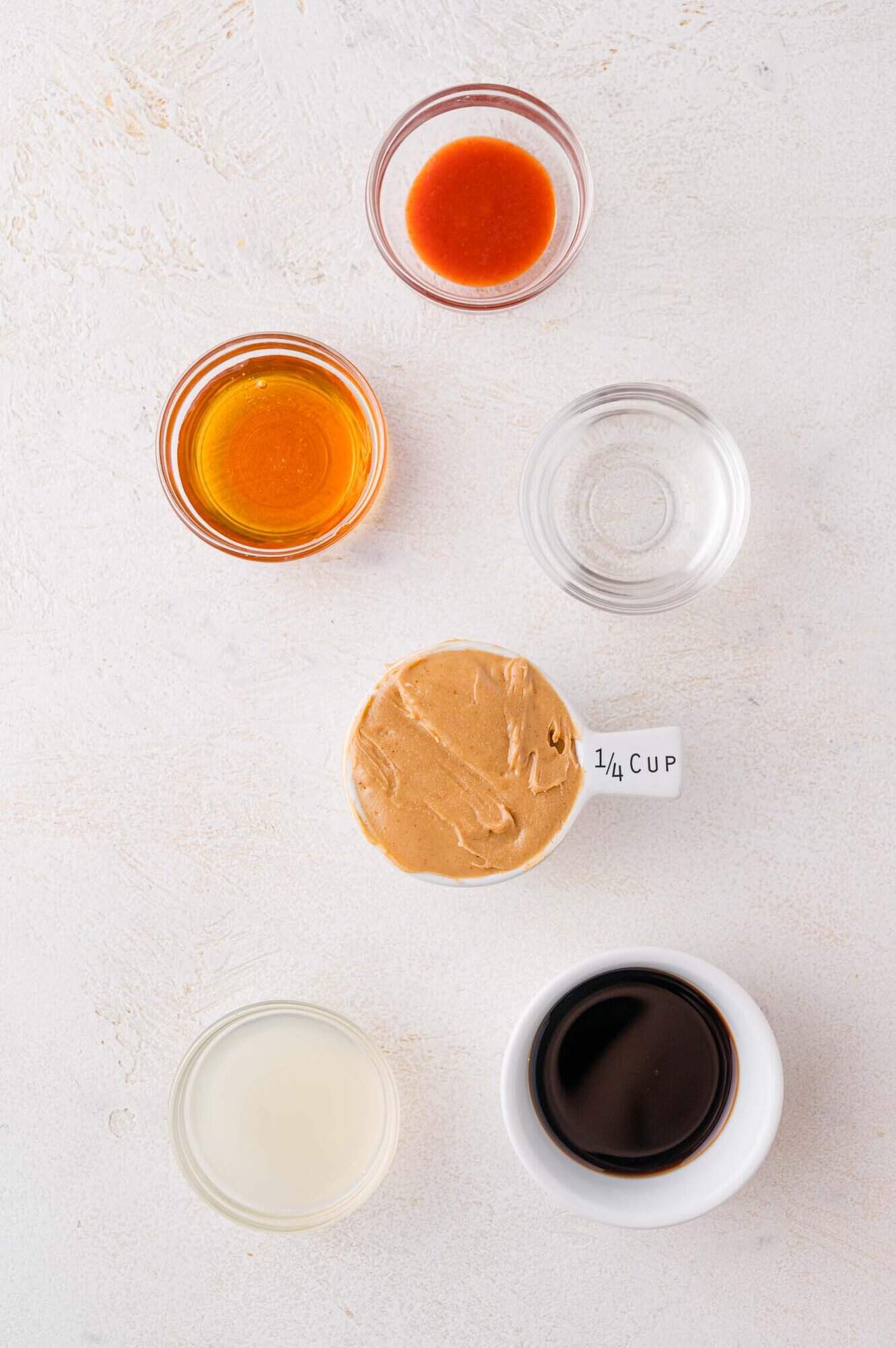 Six small bowls containing srirachia, honey, water, peanut butter, lime juice, and soy sauce arranged on a white surface.