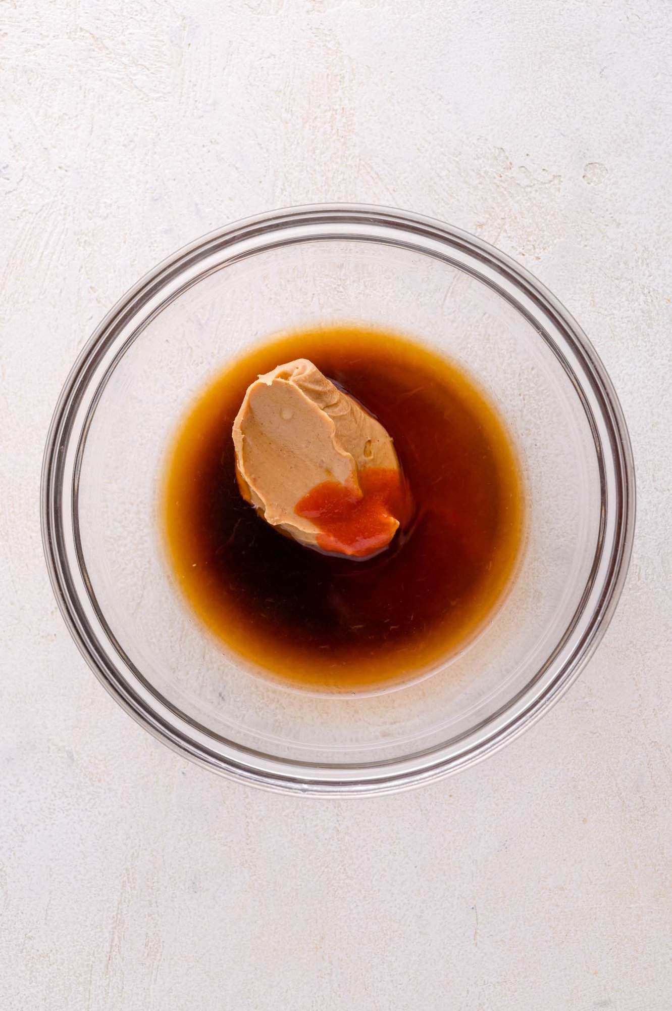 A clear glass bowl containing a scoop of peanut butter and a dark brown liquid, viewed from above on a light surface.