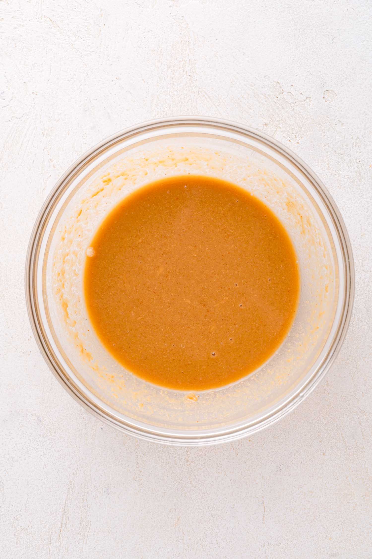 A glass bowl containing a light brown, smooth batter or sauce on a white surface.