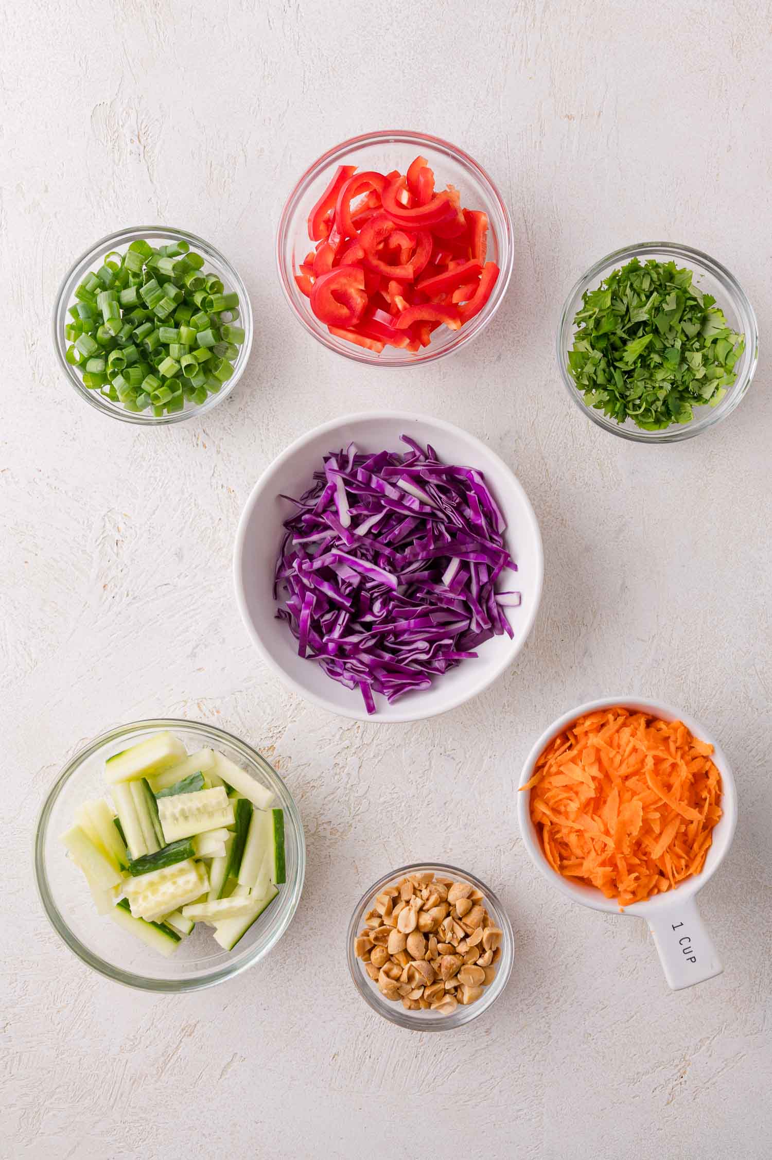 Assorted chopped vegetables and peanuts in small glass bowls, including shredded carrots, red cabbage, cucumber, green onions, red bell pepper, cilantro, and peanuts on a light background.