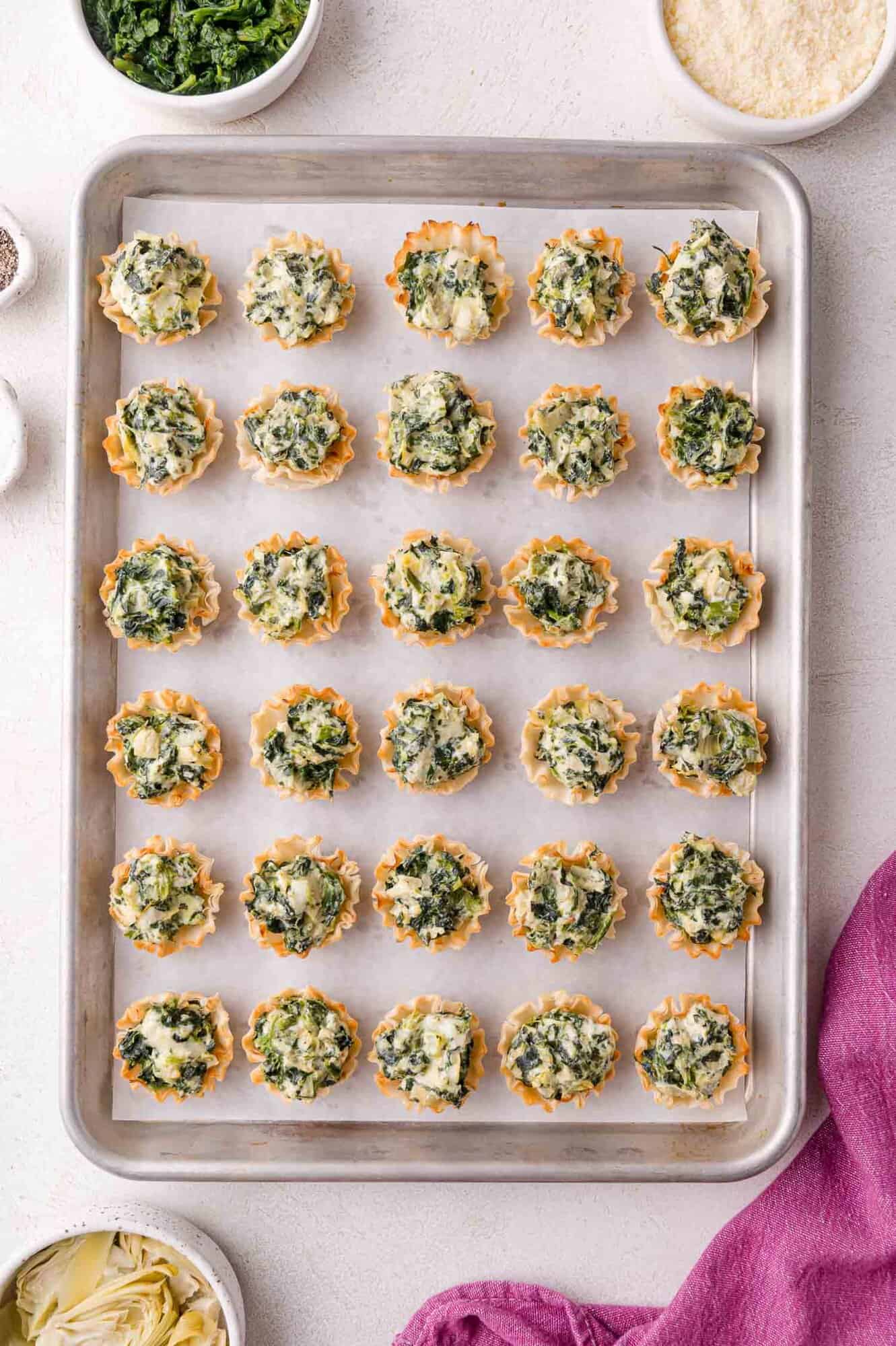 A baking sheet with 24 mini phyllo cups filled with a spinach and cheese mixture, arranged in rows on parchment paper. Bowls of ingredients are nearby.