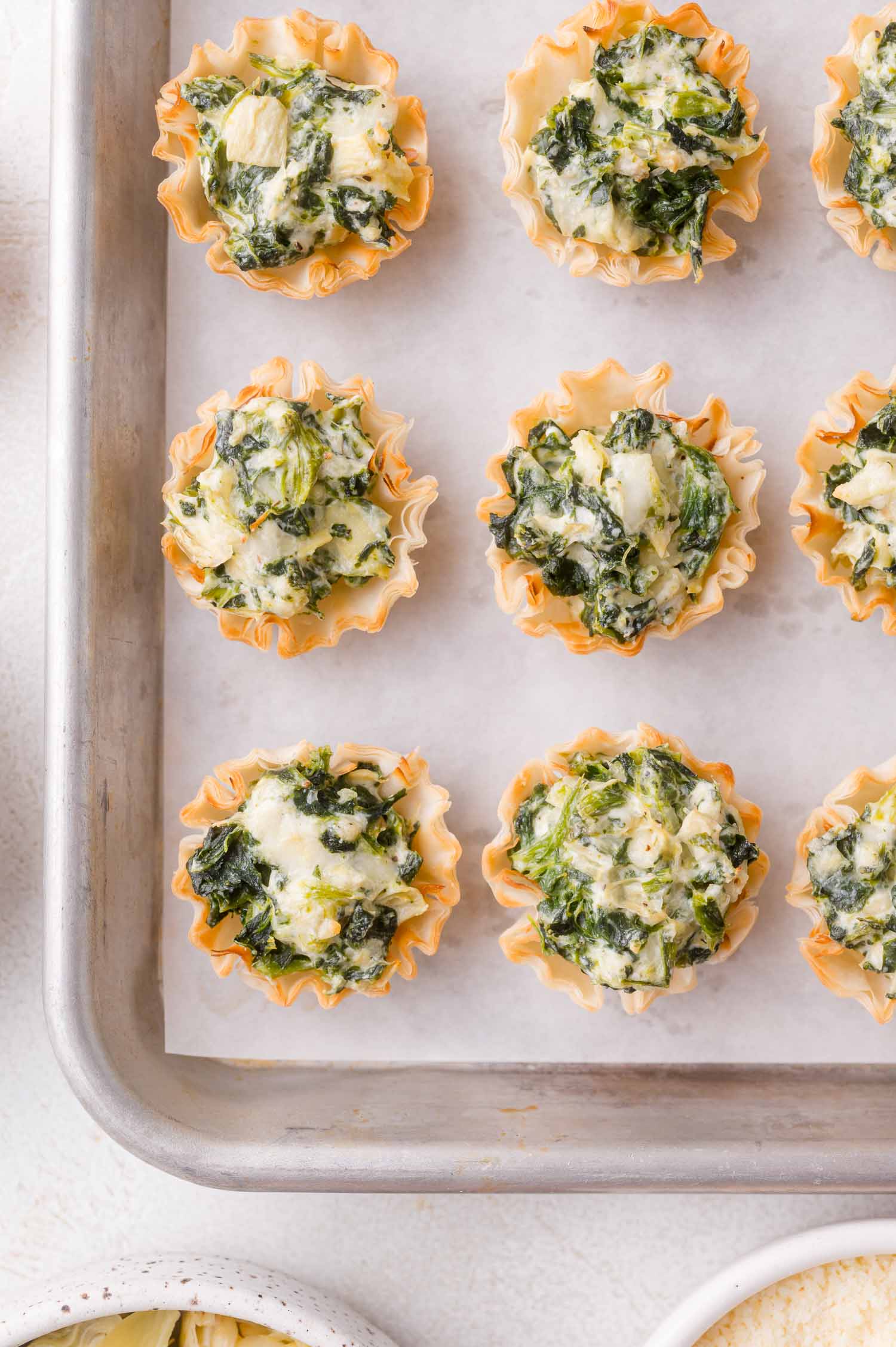Mini phyllo cups filled with spinach and artichoke dip are arranged on a parchment-lined baking sheet.