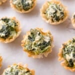 Mini phyllo pastry cups filled with a creamy spinach mixture are arranged on a parchment-lined surface.