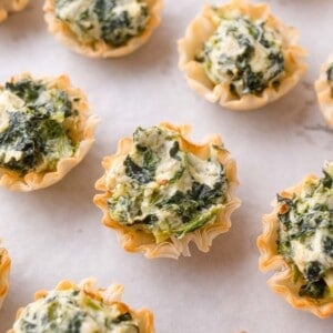 Mini phyllo pastry cups filled with a creamy spinach mixture are arranged on a parchment-lined surface.