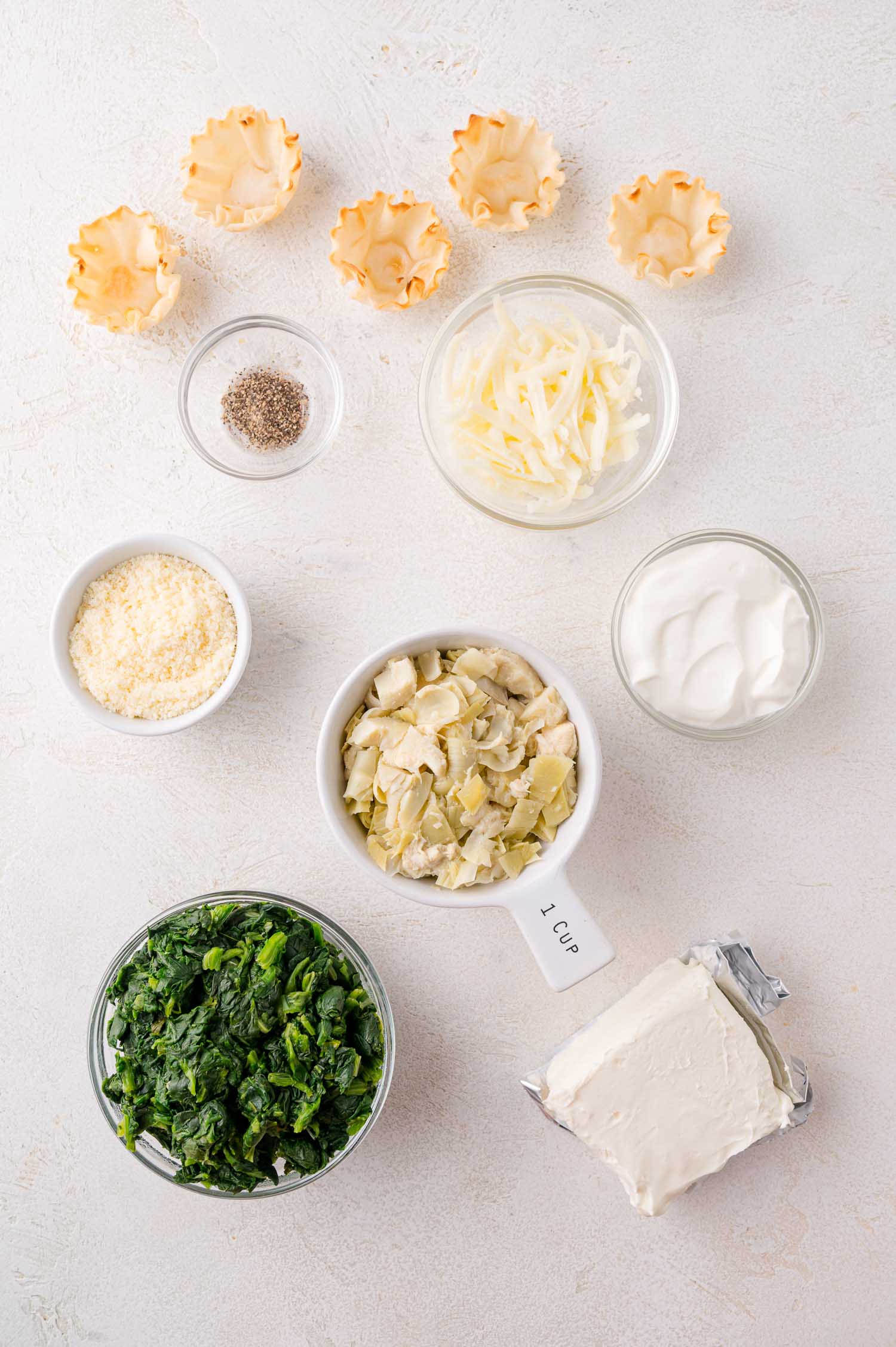 Ingredients for spinach artichoke bites arranged on a white surface, including mini phyllo shells, shredded cheese, parmesan, artichoke hearts, spinach, cream cheese, sour cream, and pepper.