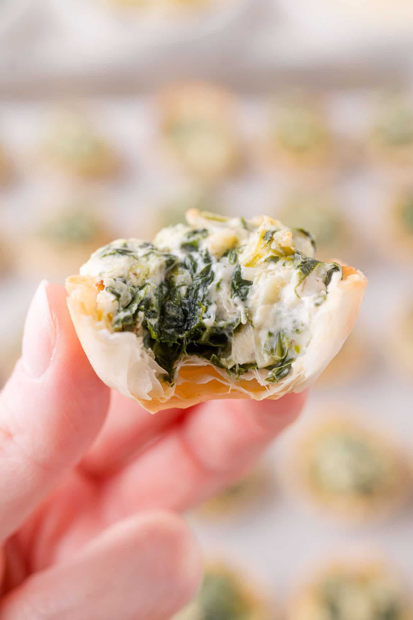 A hand holding a small phyllo pastry cup filled with spinach and cheese filling, with a bite taken out to show the inside.