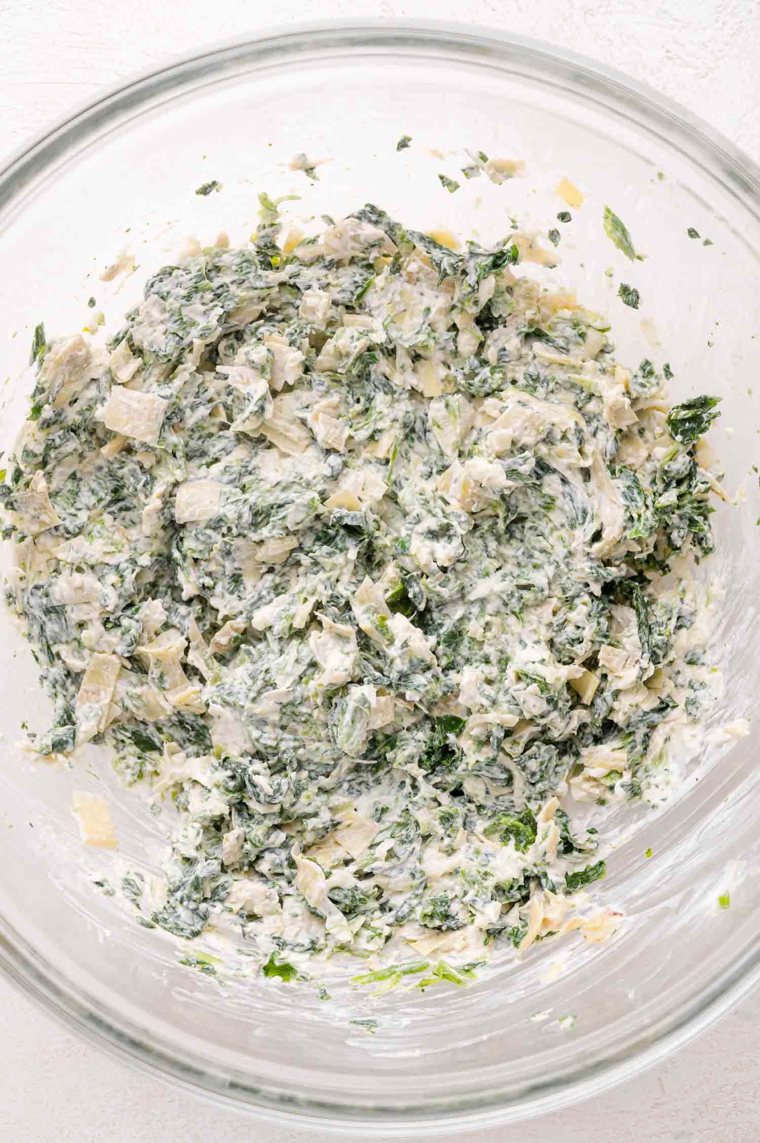 A glass bowl containing a mixture of chopped spinach, artichokes, and creamy white cheese.