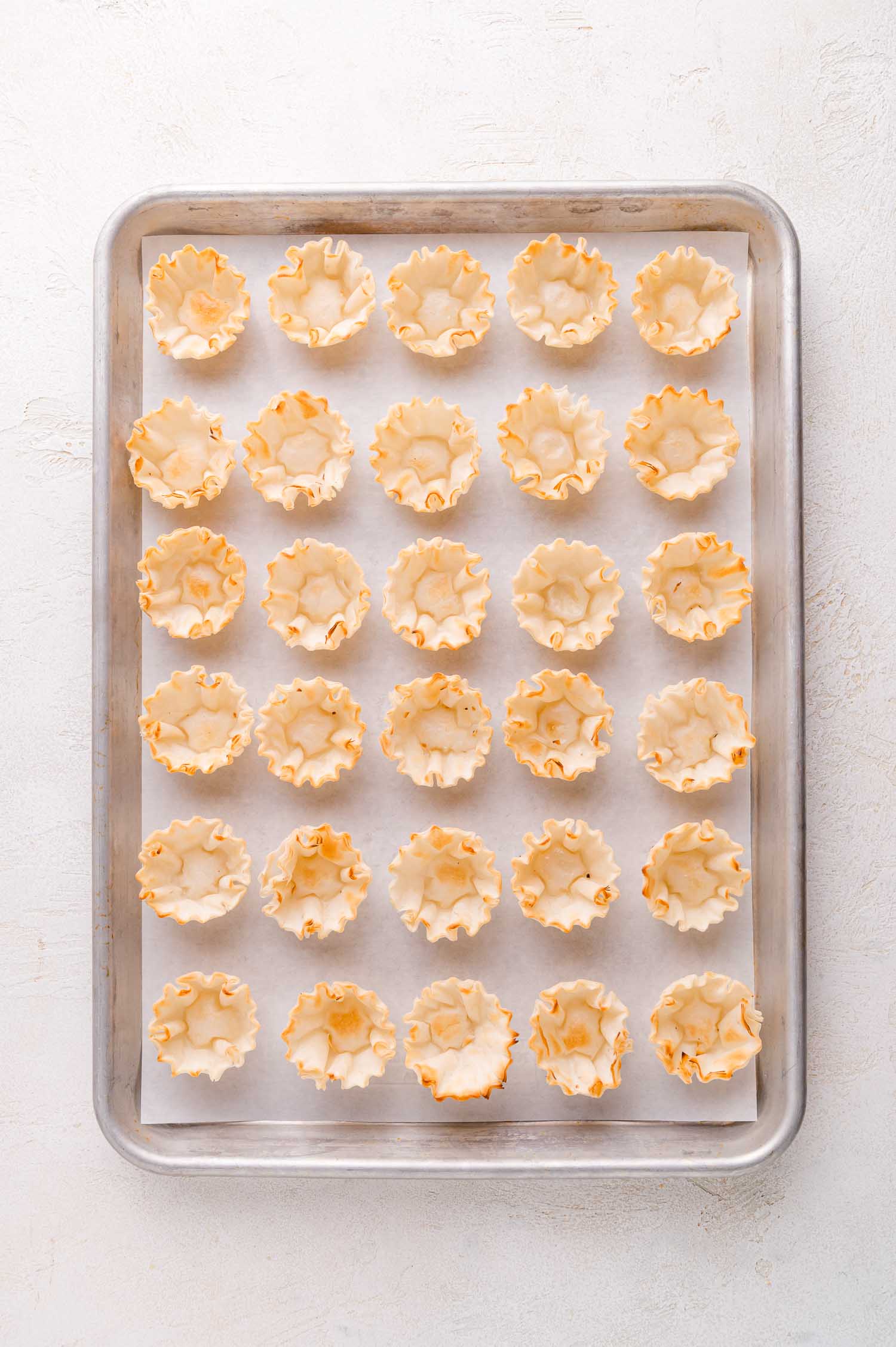 A baking tray lined with parchment paper holds 30 empty mini phyllo tart shells arranged in a grid pattern.