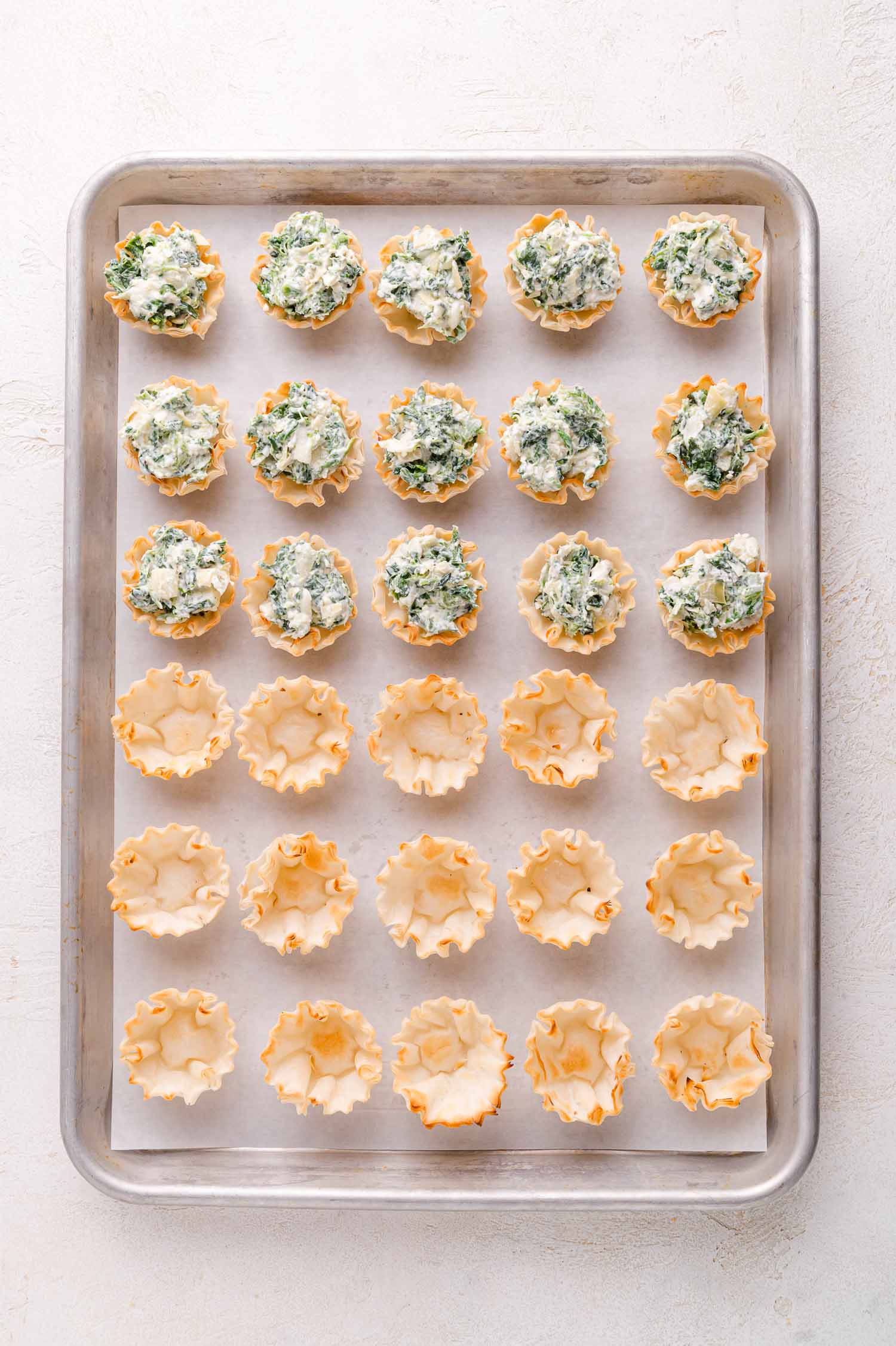 A baking sheet with rows of empty phyllo pastry cups on the bottom half and spinach-cheese filled pastry cups on the top half, arranged on parchment paper.