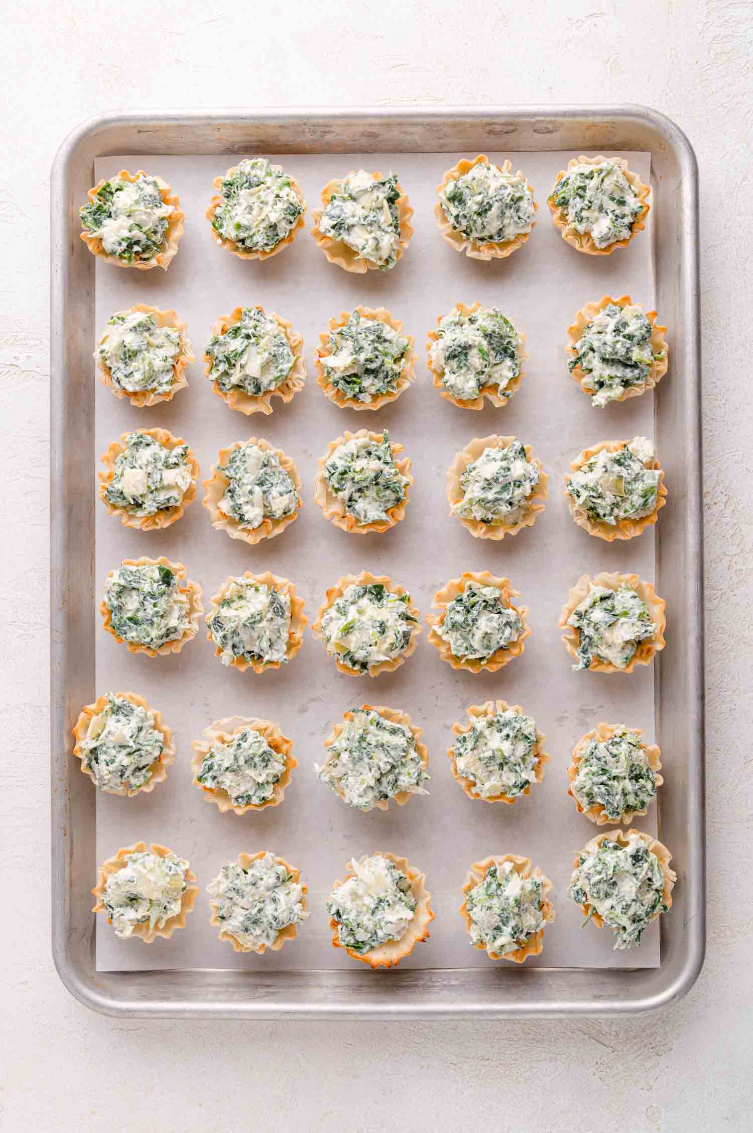A baking sheet with neatly arranged rows of mini phyllo pastry cups filled with a creamy spinach and cheese mixture.