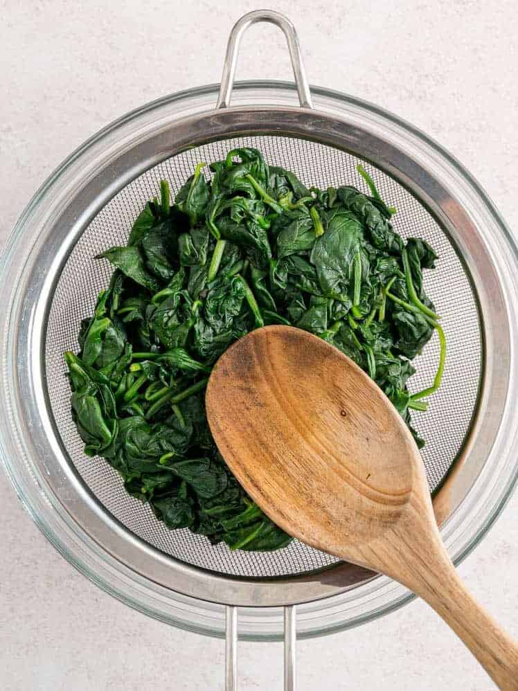 A wooden spoon pressed wilted spinach against a fine mesh strainer to drain the excess liquid.