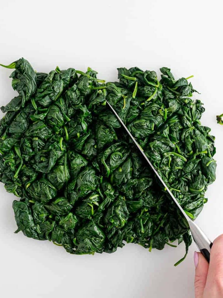 A hand uses a knife to chop cooked spinach on a white surface.