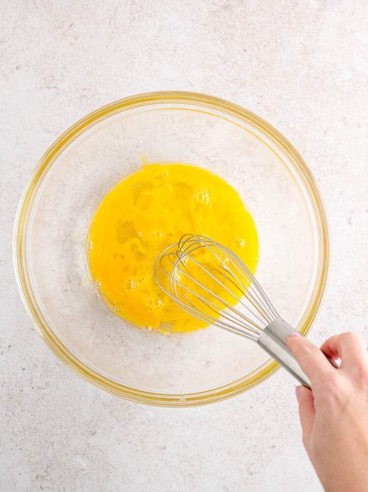 A hand uses a whisk to beat eggs in a glass bowl.