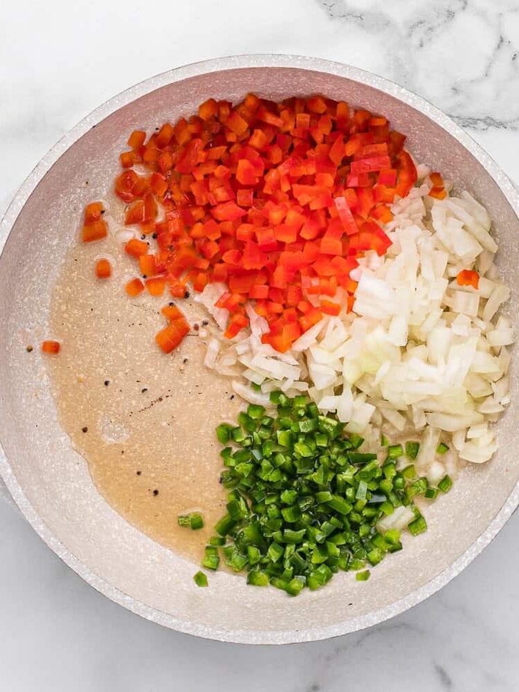 Chopped red bell pepper, onion, and jalapeño in a white pan with oil, ready to be sautéed, on a marble surface.