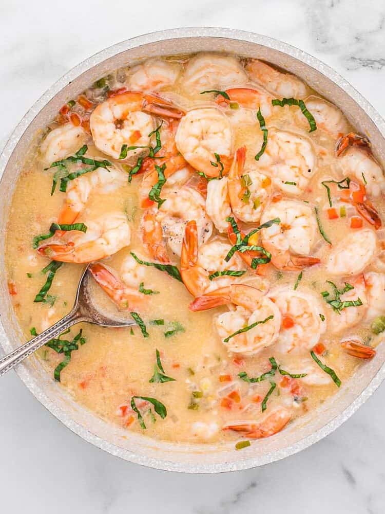 A pot of shrimp in a creamy sauce with chopped herbs and diced vegetables, served with a metal spoon on a marble surface.