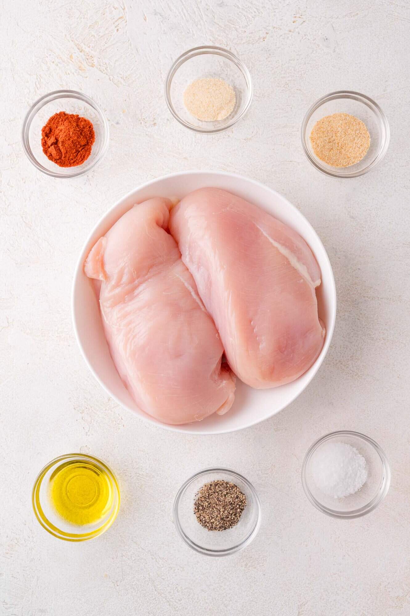 Raw chicken breasts in a white bowl surrounded by small bowls of olive oil, black pepper, salt, paprika, garlic powder, and onion powder on a white surface.