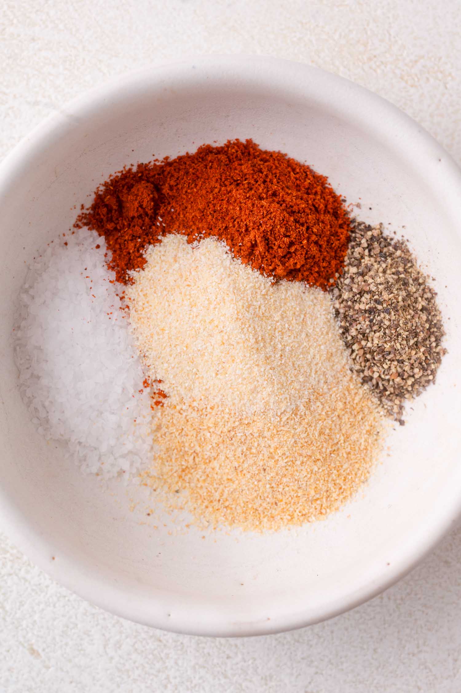A white bowl containing separate piles of salt, paprika, ground black pepper, garlic powder, and onion powder.