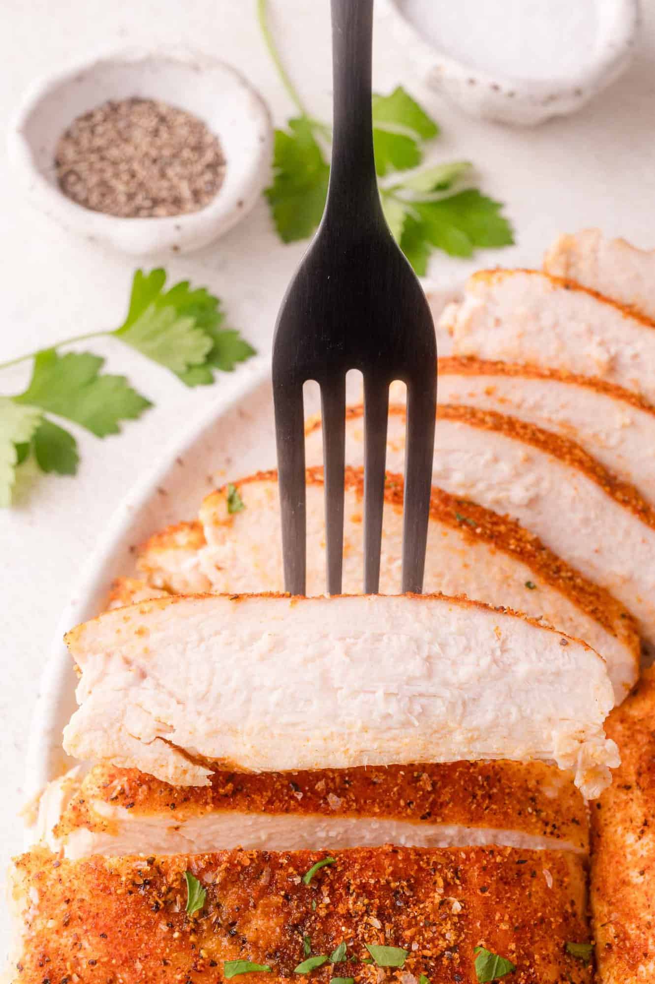 A black fork holds a slice of seasoned, cooked chicken breast on a white plate with herbs, salt, and pepper in the background.