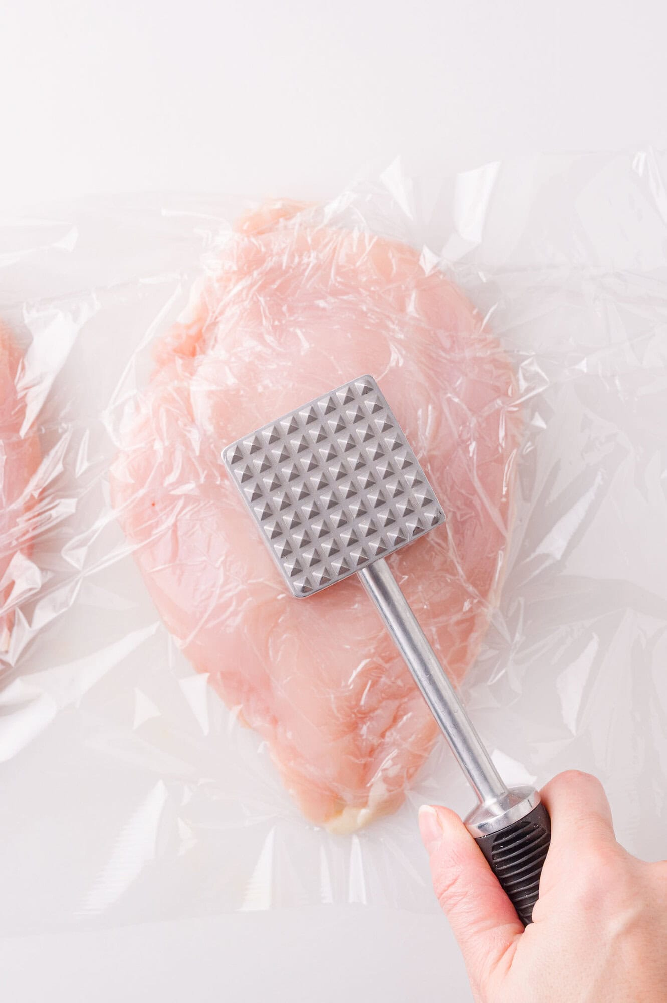 A hand uses a metal meat tenderizer to pound a raw chicken breast covered with plastic wrap on a white surface.