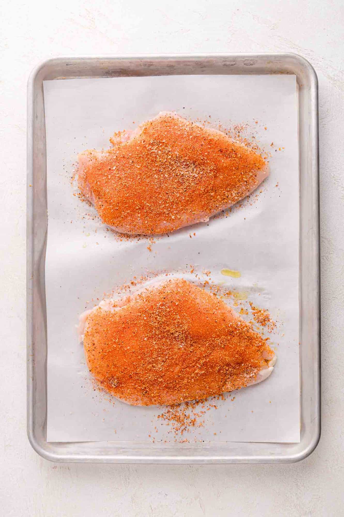 Two raw chicken breasts on a parchment-lined baking sheet, coated with a reddish-brown spice rub.