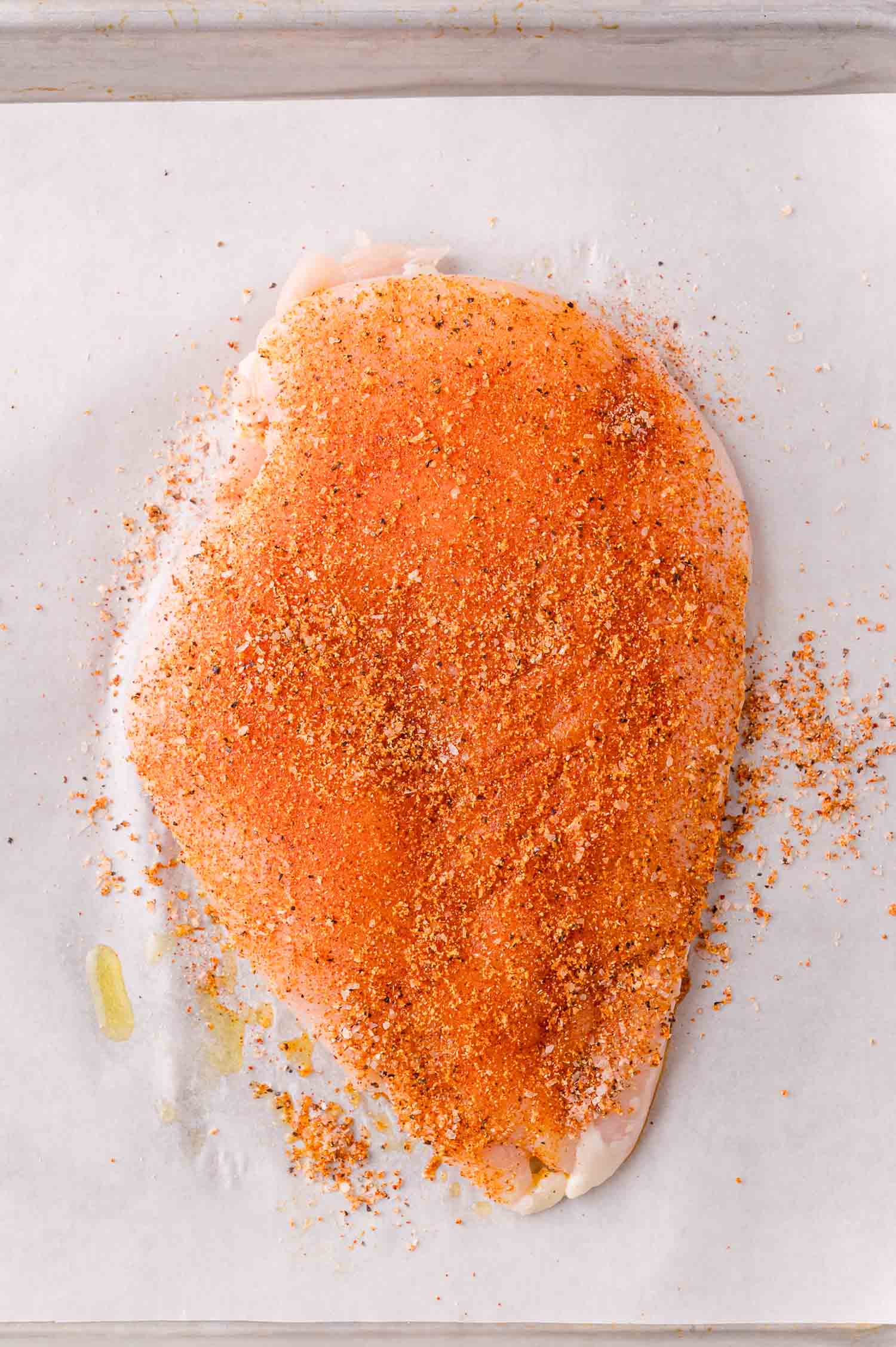Raw chicken breast covered with a layer of red and brown seasoning mix on a sheet of parchment paper.
