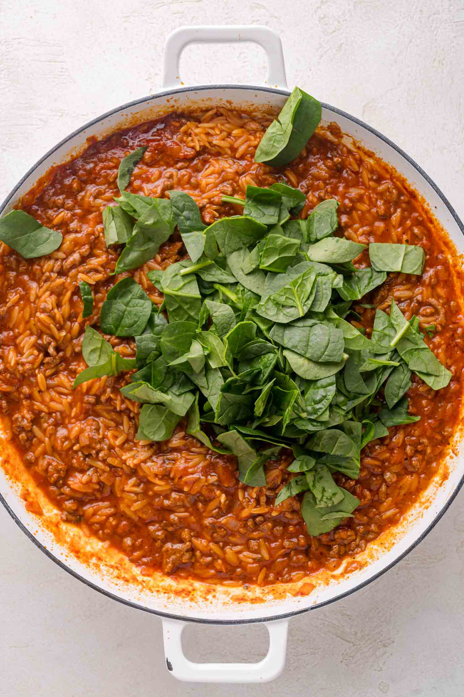 A white pot filled with tomato-based orzo pasta topped with fresh spinach leaves on a light surface.