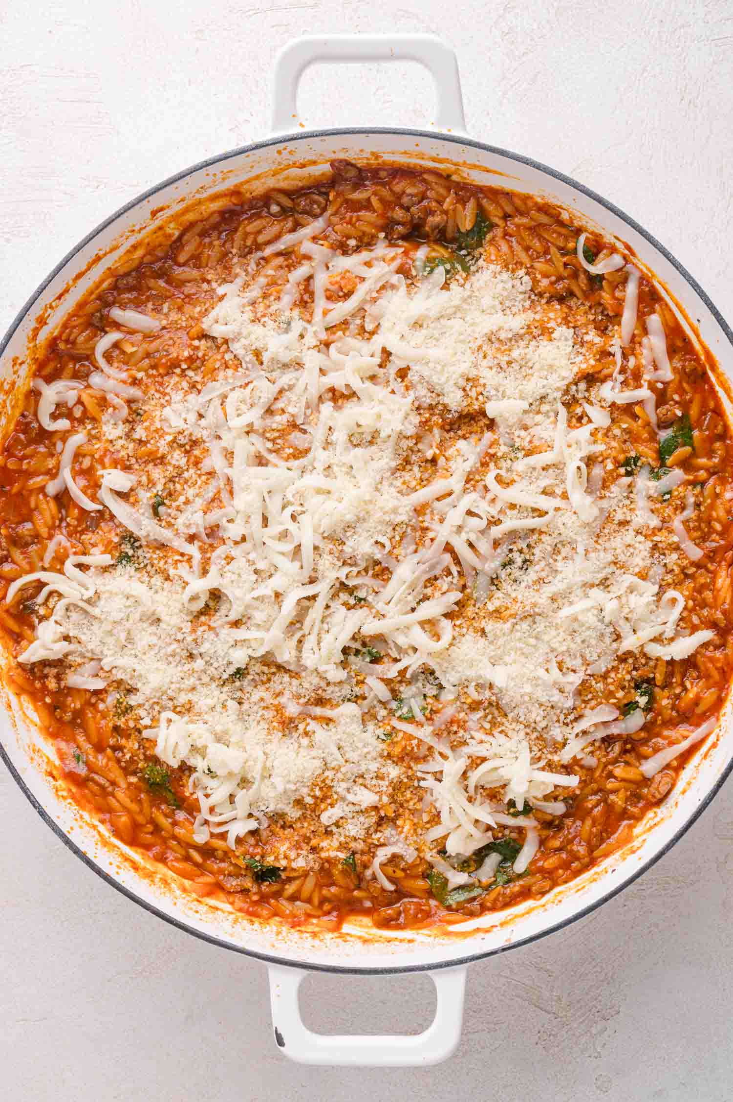A white pot filled with tomato-based orzo pasta topped with shredded mozzarella and grated parmesan cheese.