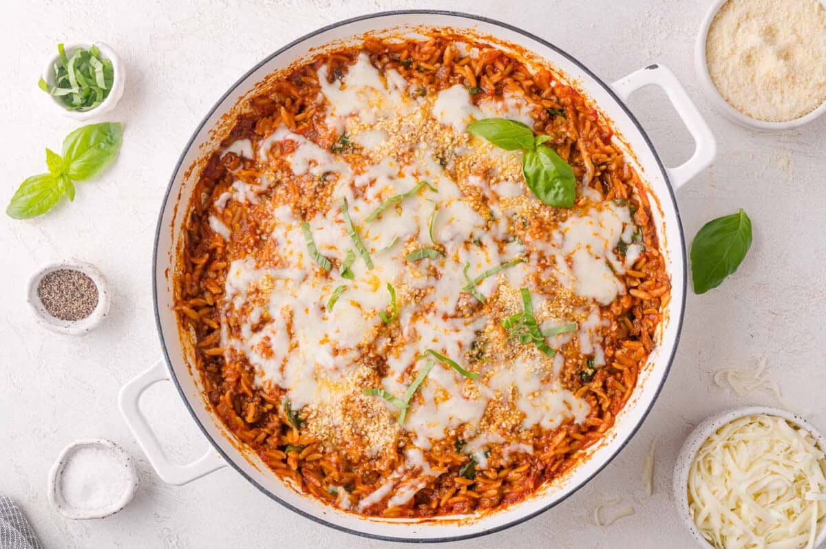 A white casserole dish filled with baked orzo pasta in tomato sauce, topped with melted cheese and garnished with fresh basil, surrounded by small bowls of cheese, pepper, and herbs.