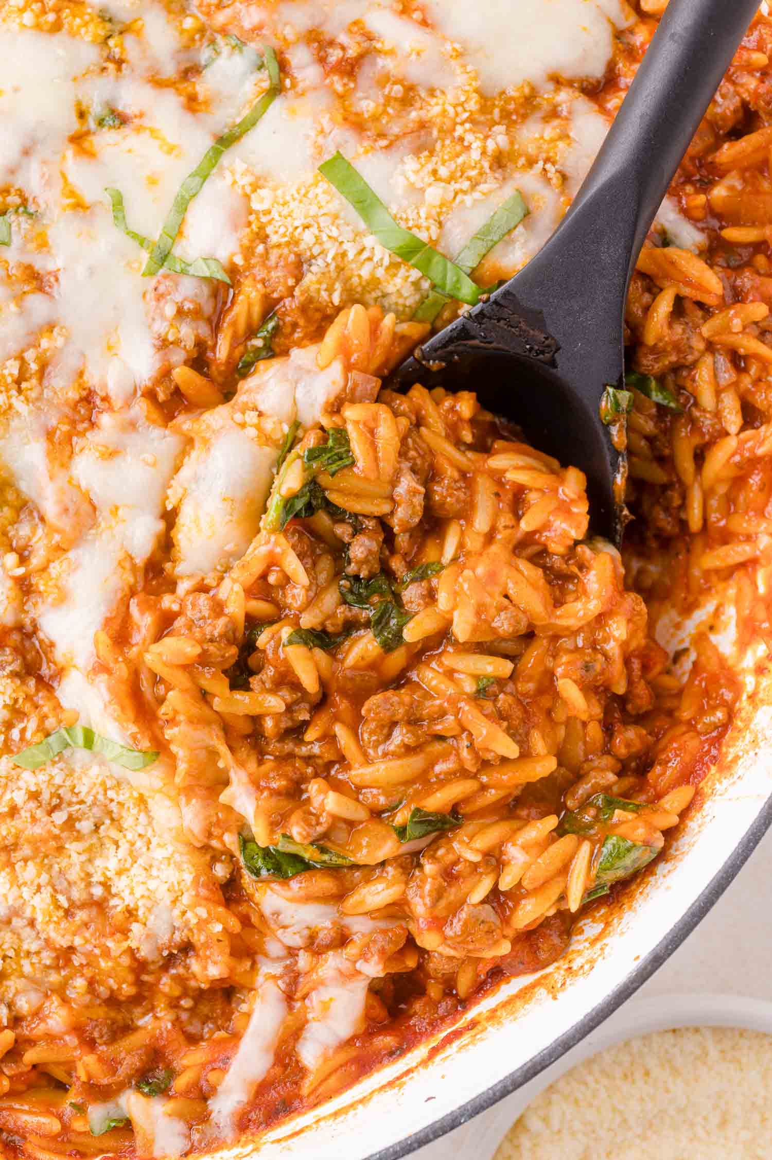 A close-up of a baked orzo pasta dish with ground meat, tomato sauce, melted cheese, and fresh basil, being served with a black spoon.