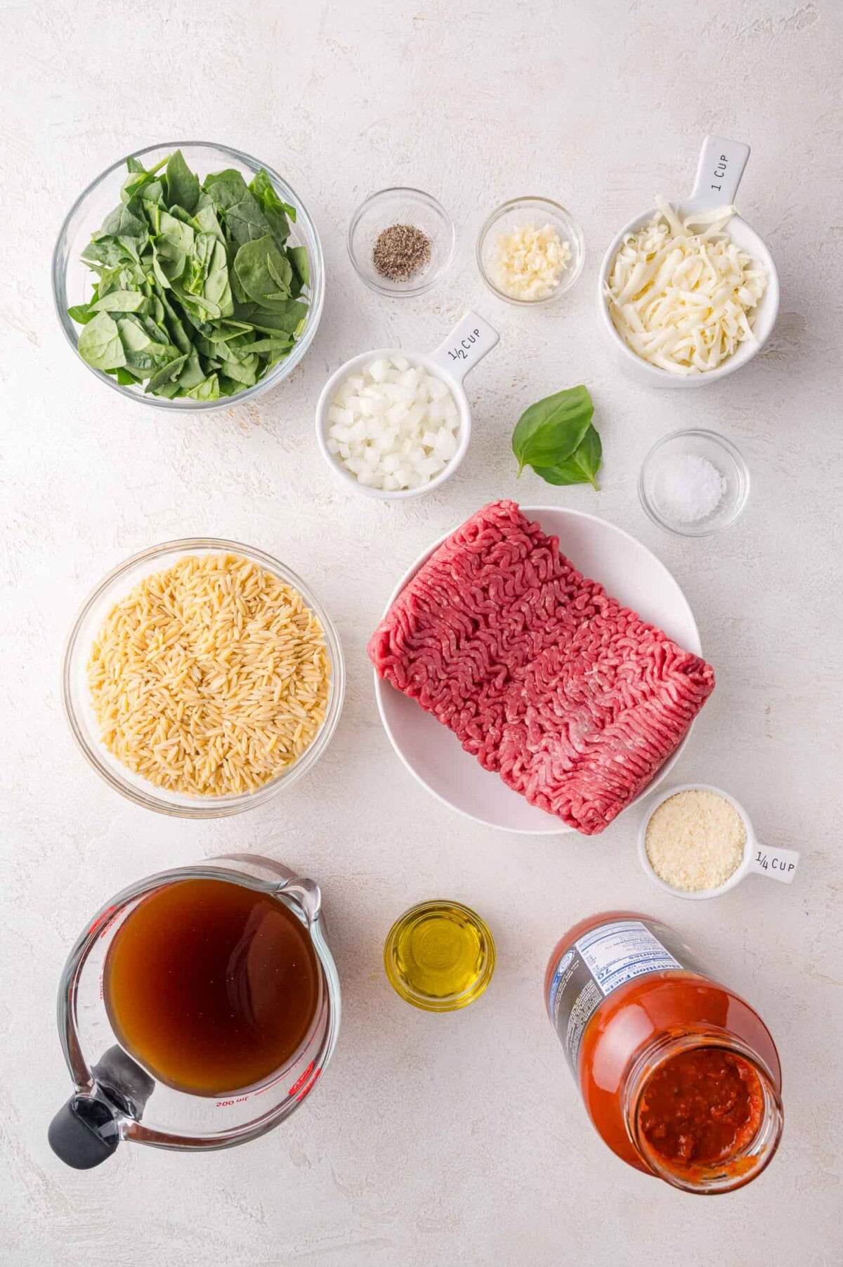 Ingredients for a pasta dish arranged on a white surface, including ground beef, orzo, spinach, cheese, onions, garlic, olive oil, broth, marinara sauce, and seasonings in bowls and measuring cups.