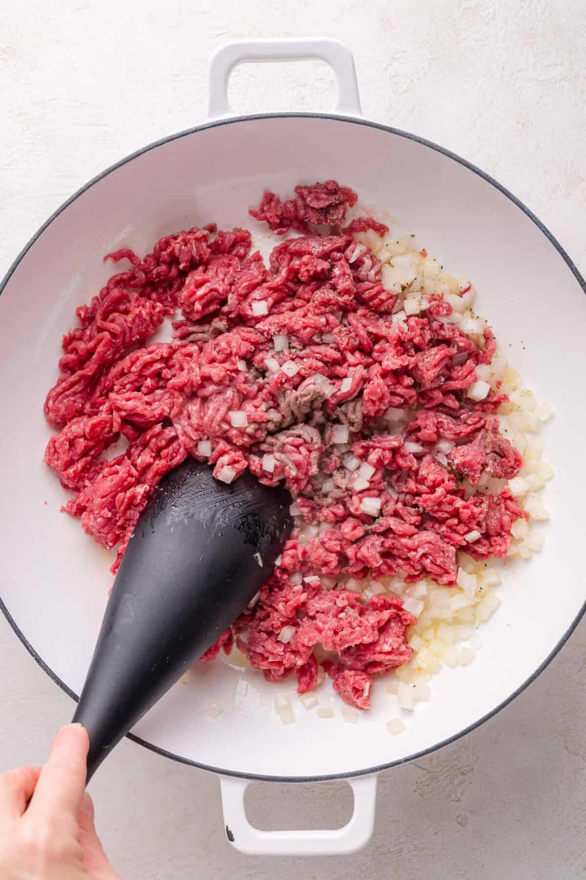 Ground beef and chopped onions being sautéed in a white pan, stirred with a black spatula.