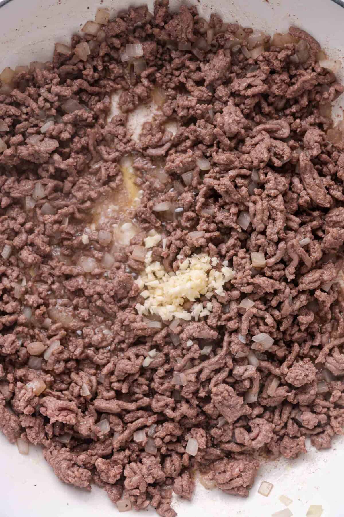 Cooked ground beef and diced onions in a pan with a small pile of minced garlic added to the center.