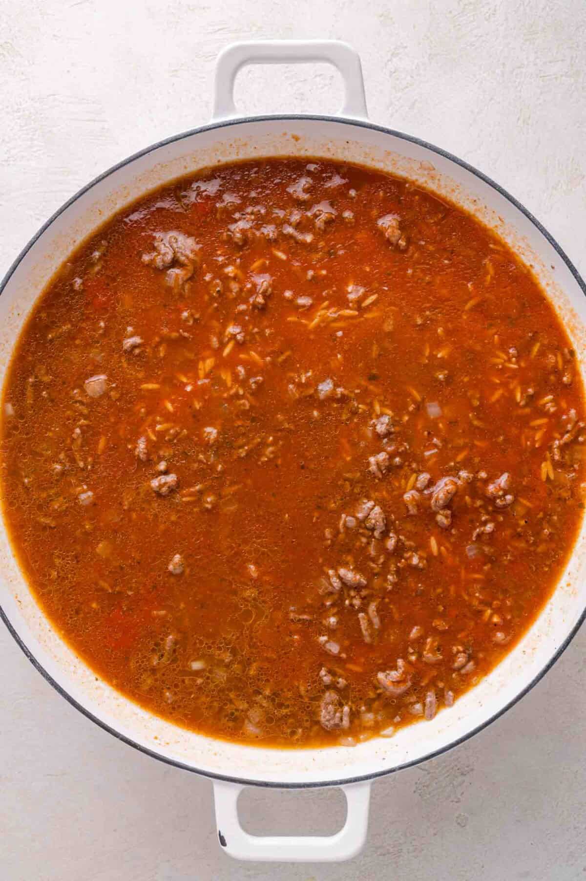 Large white pot filled with tomato-based mixture containing ground meat, orzo, and herbs, placed on a light-colored surface.