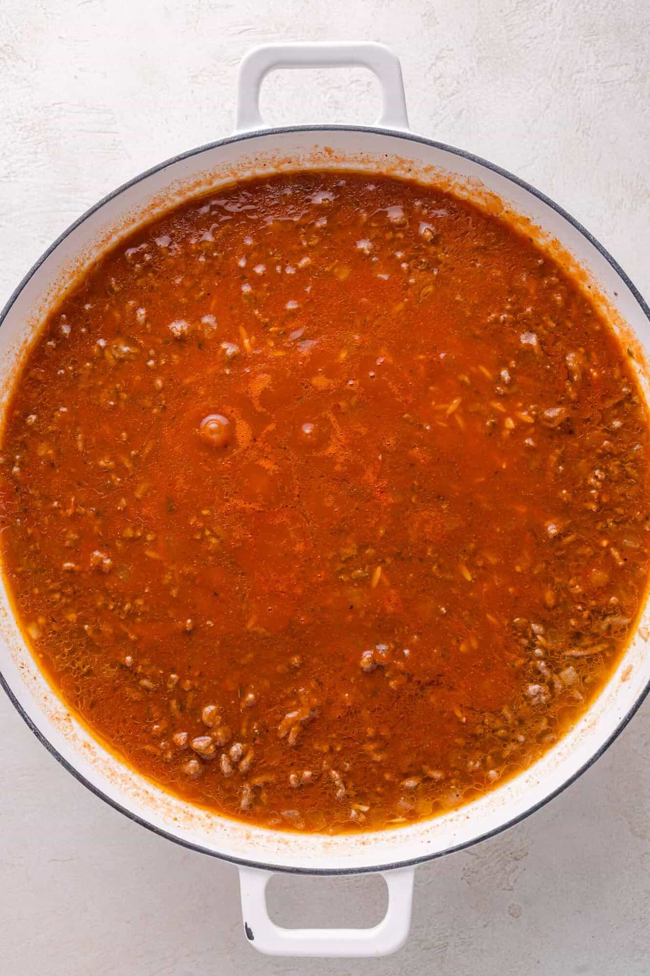 Large white pot filled with chunky red meat sauce, simmering, seen from above on a light surface.