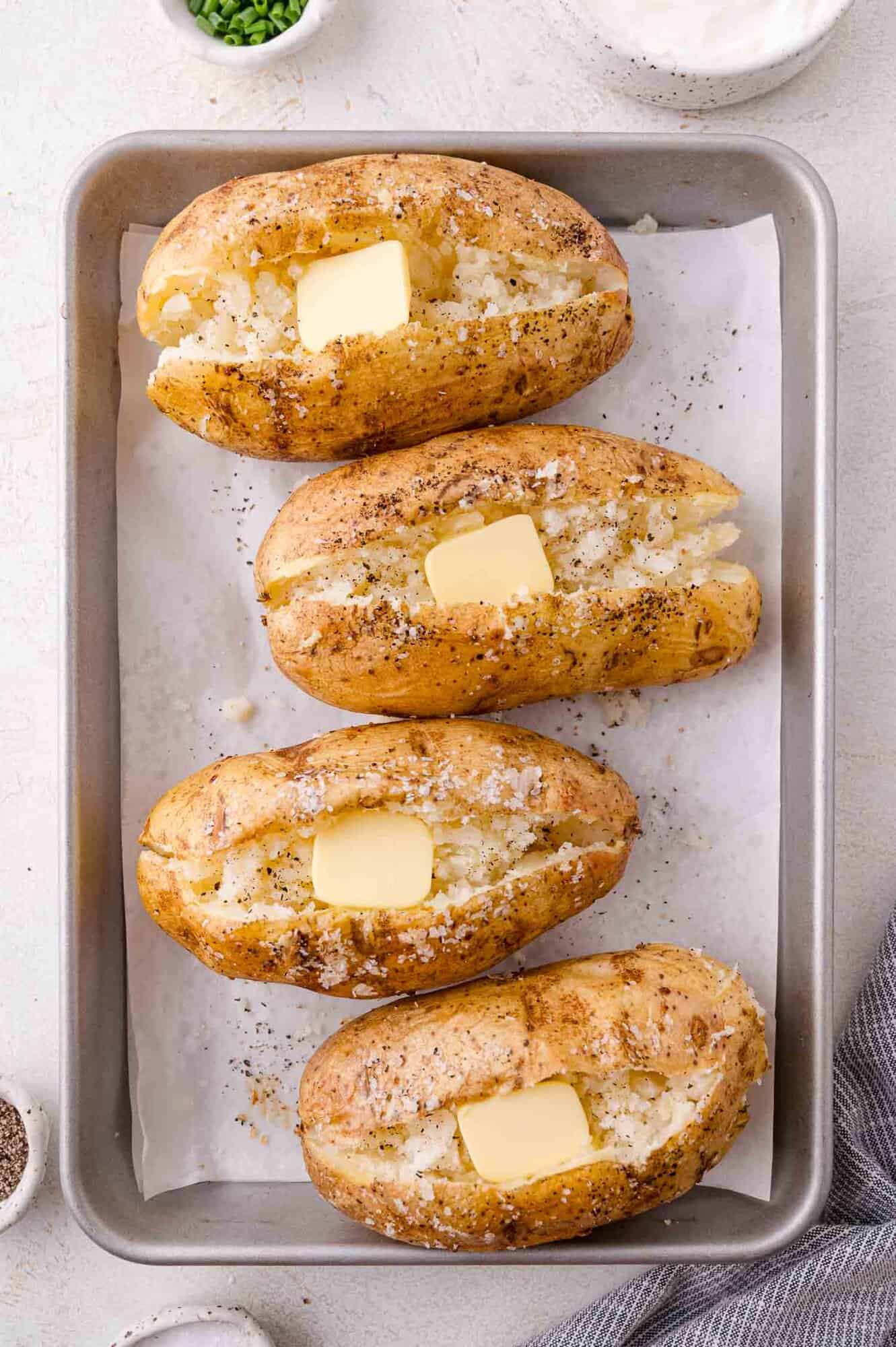 Four baked potatoes with cracked tops, each topped with a pat of butter and sprinkled with salt and pepper, arranged on a parchment-lined baking sheet.