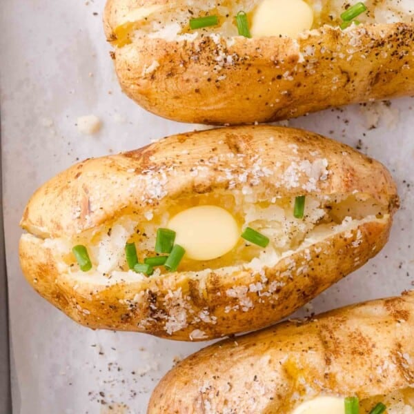 Three baked potatoes with crispy skins are split open, topped with pats of butter, chopped chives, salt, and pepper, on a parchment-lined surface.