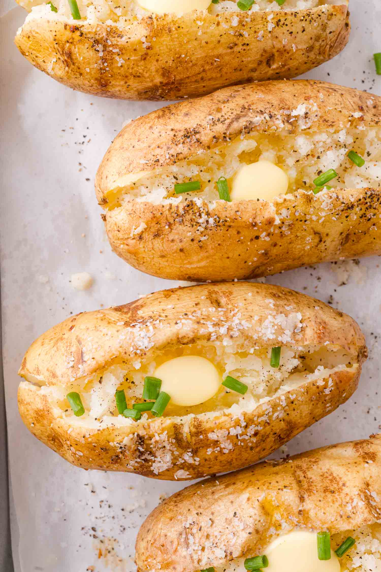 Baked potatoes topped with butter, chopped chives, and coarse salt on a parchment-lined baking sheet.