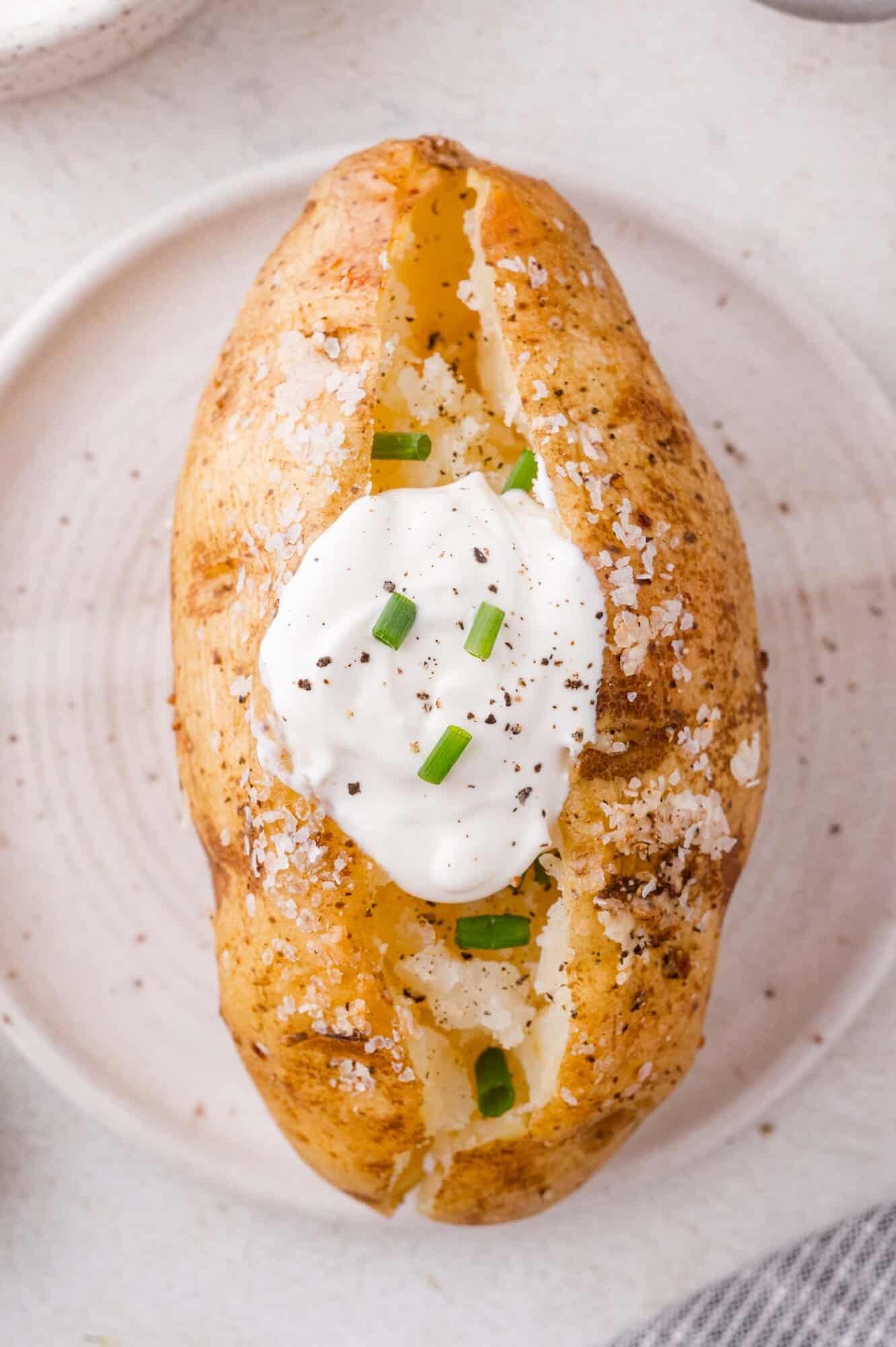 A baked potato with a cracked open top, topped with sour cream, chopped chives, black pepper, and coarse salt, served on a white plate.
