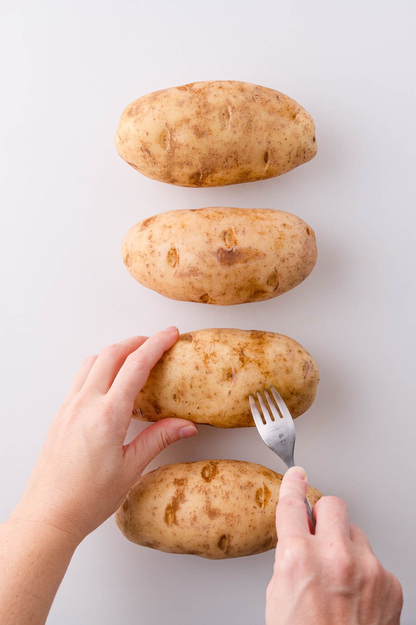 Four russet potatoes on a white surface, with a hand using a fork to poke holes in one of the potatoes.