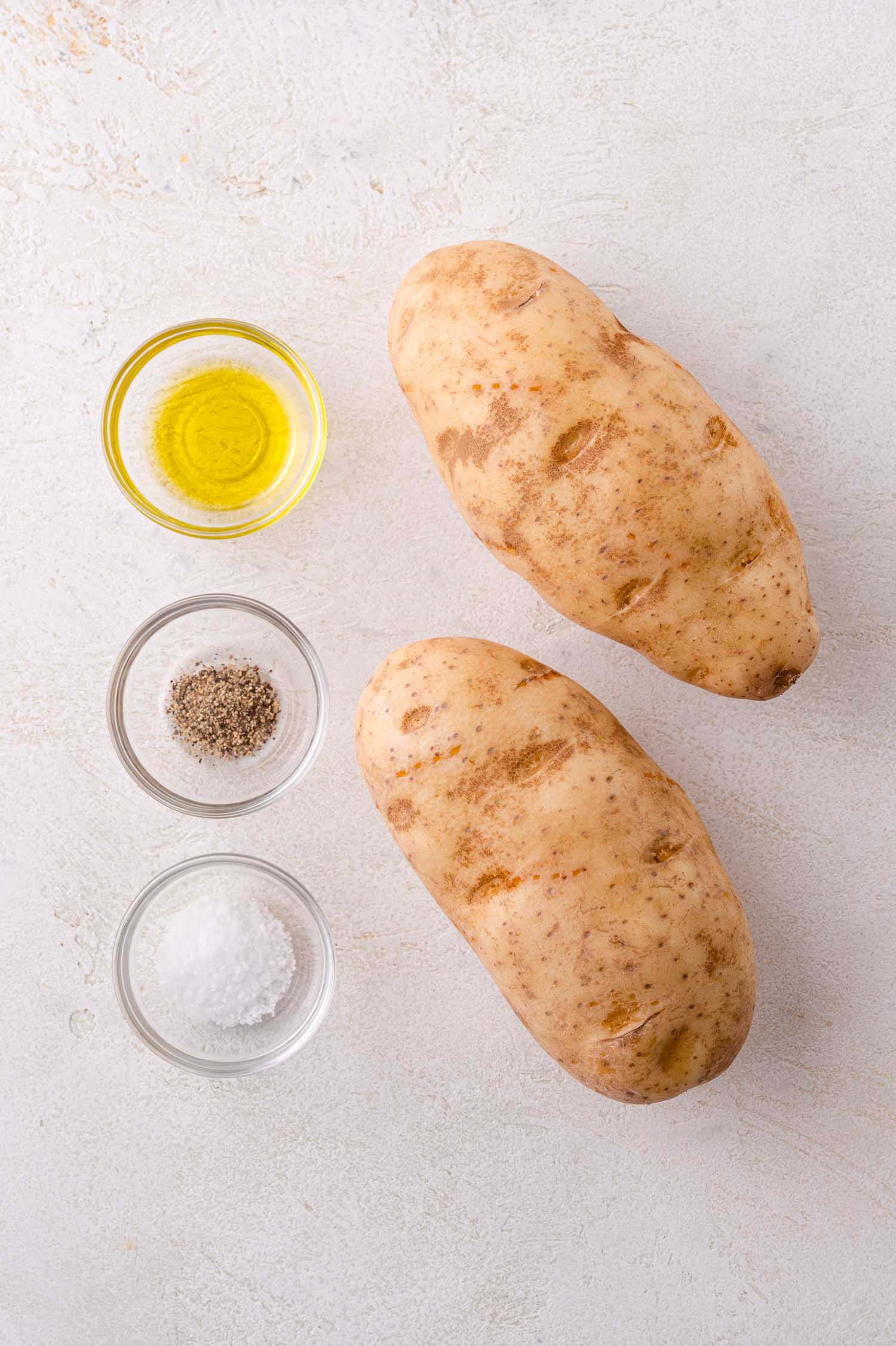 Two whole russet potatoes on a light surface, with small glass bowls of olive oil, ground black pepper, and coarse salt arranged beside them.