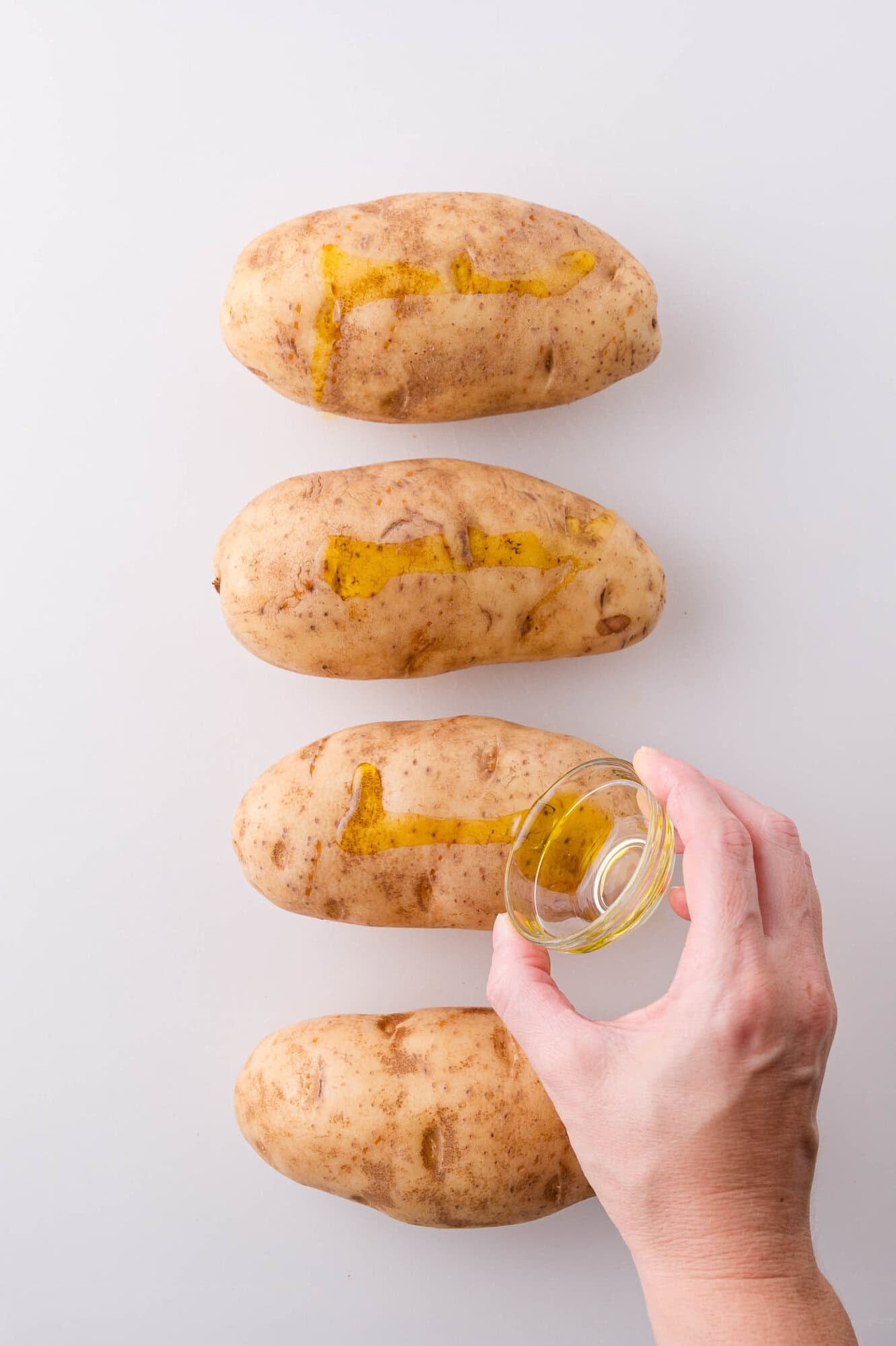 Four whole potatoes are placed in a row on a white surface, with oil being poured onto one by a hand holding a small glass bowl.