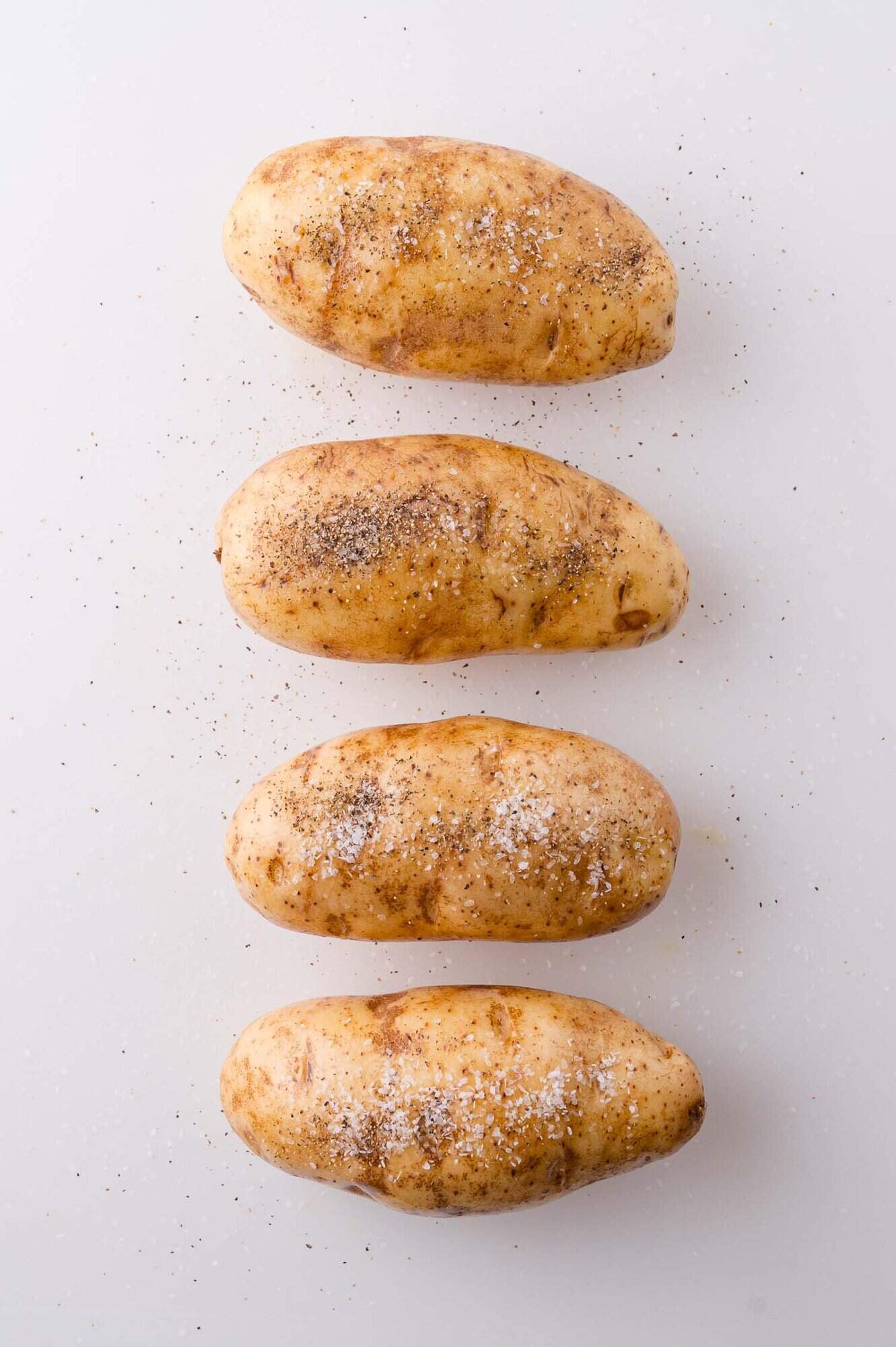 Four whole russet potatoes on a white surface, each sprinkled with salt and black pepper.