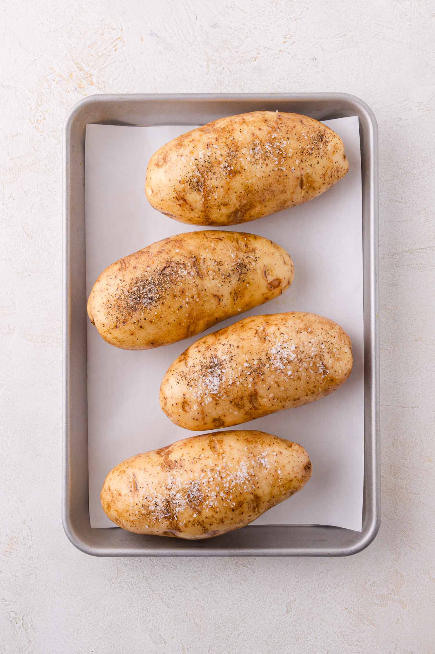 Four whole, seasoned potatoes arranged on a parchment-lined baking sheet, ready to be baked.