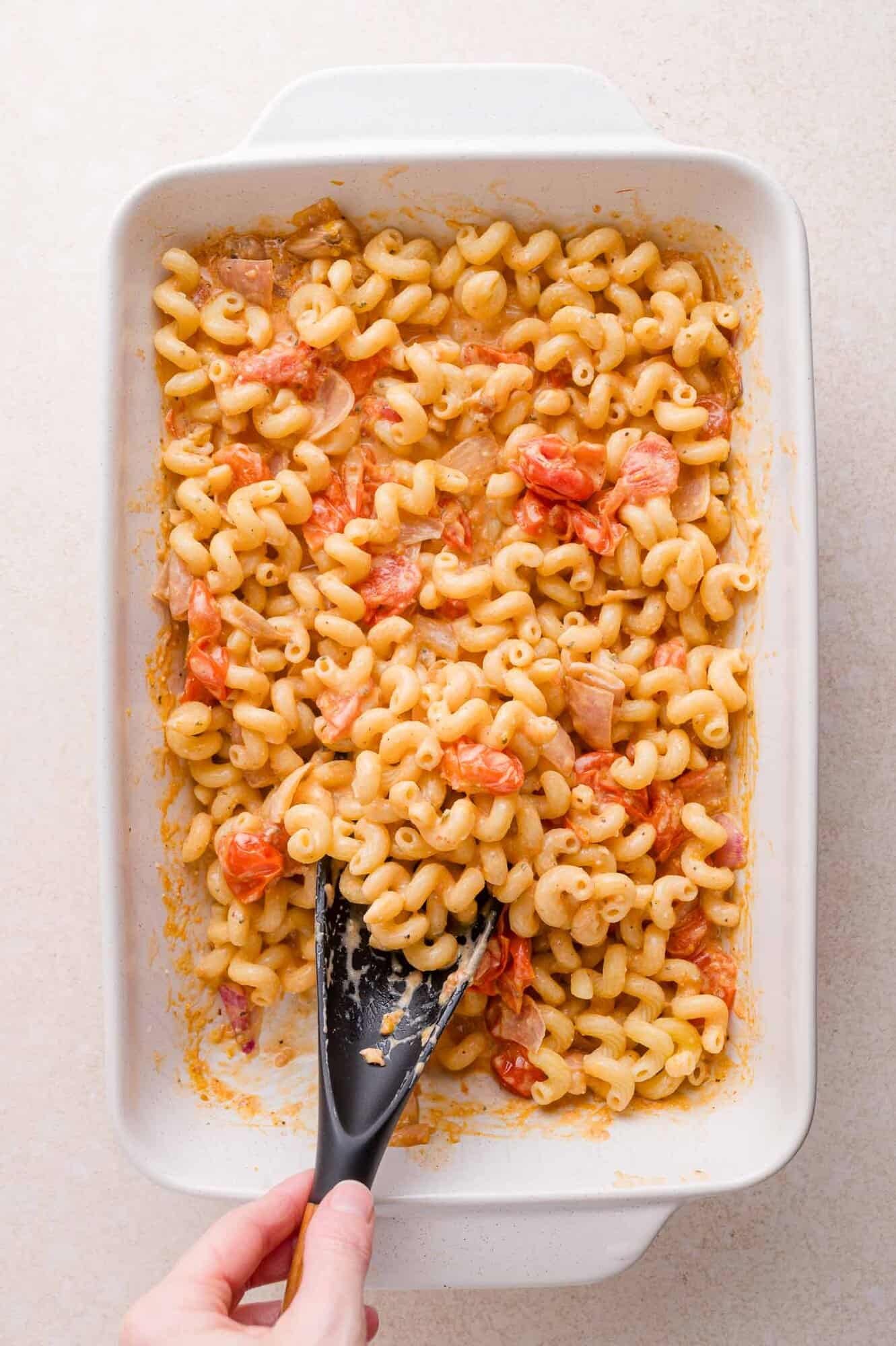 Pasta with tomatoes and sauce in a white dish, being stirred with a black spatula by a hand.