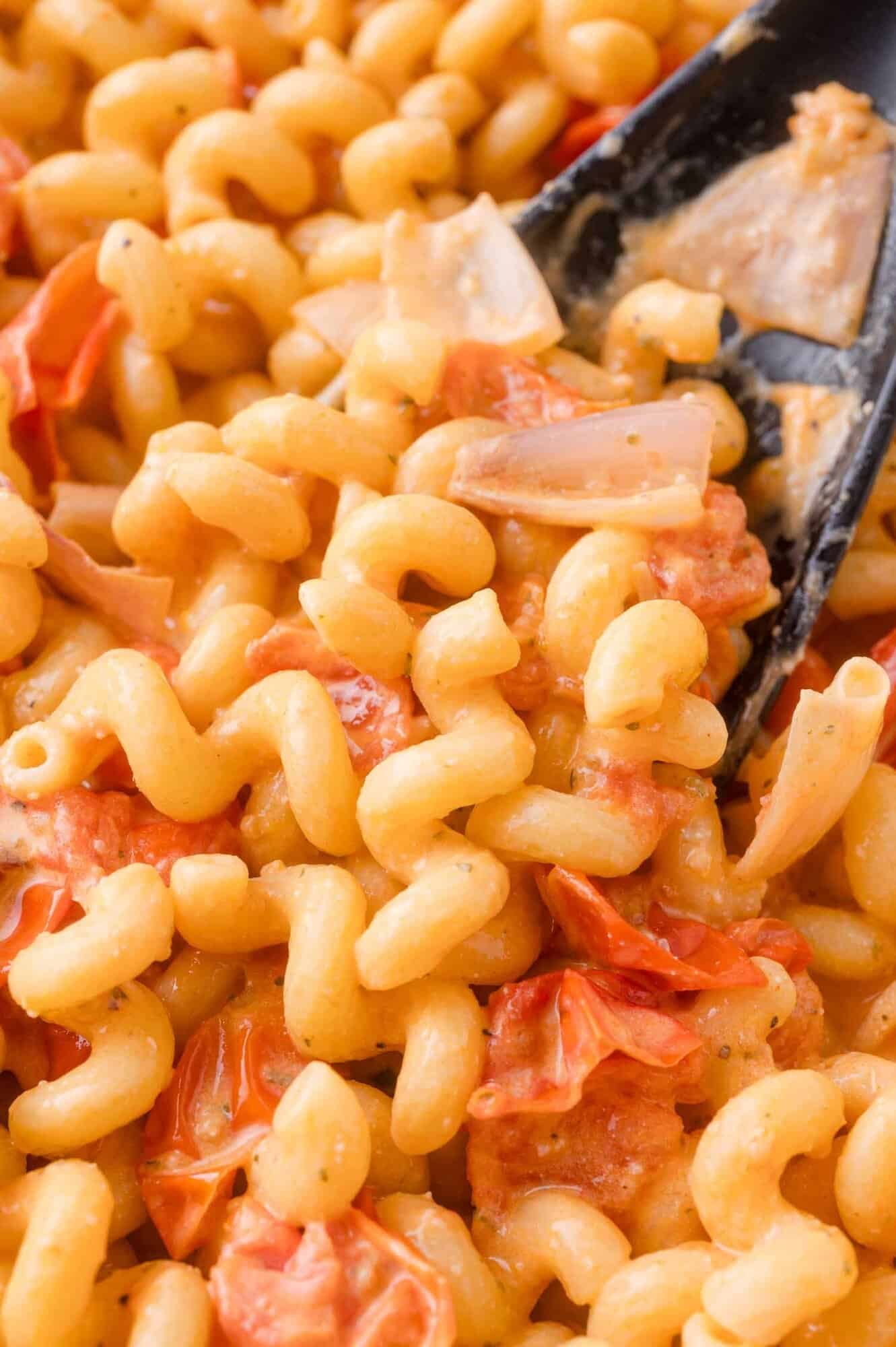 Close-up of cooked cavatappi pasta mixed with creamy sauce, tomato pieces, and sliced onions, with a serving spoon partially visible.