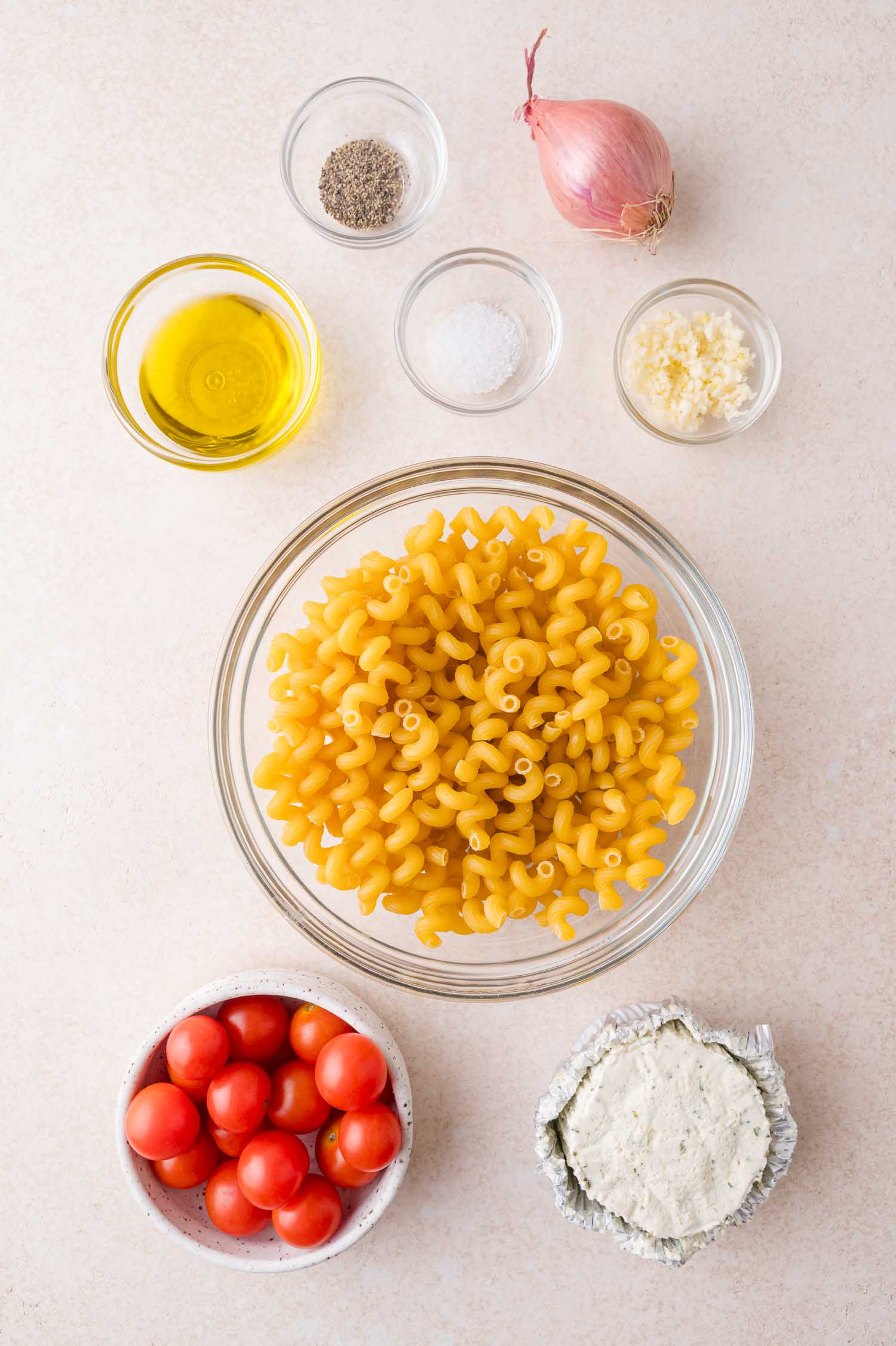 A flat lay of uncooked pasta, cherry tomatoes, boursin cheese, olive oil, minced garlic, salt, pepper, and a shallot in separate bowls on a light surface.